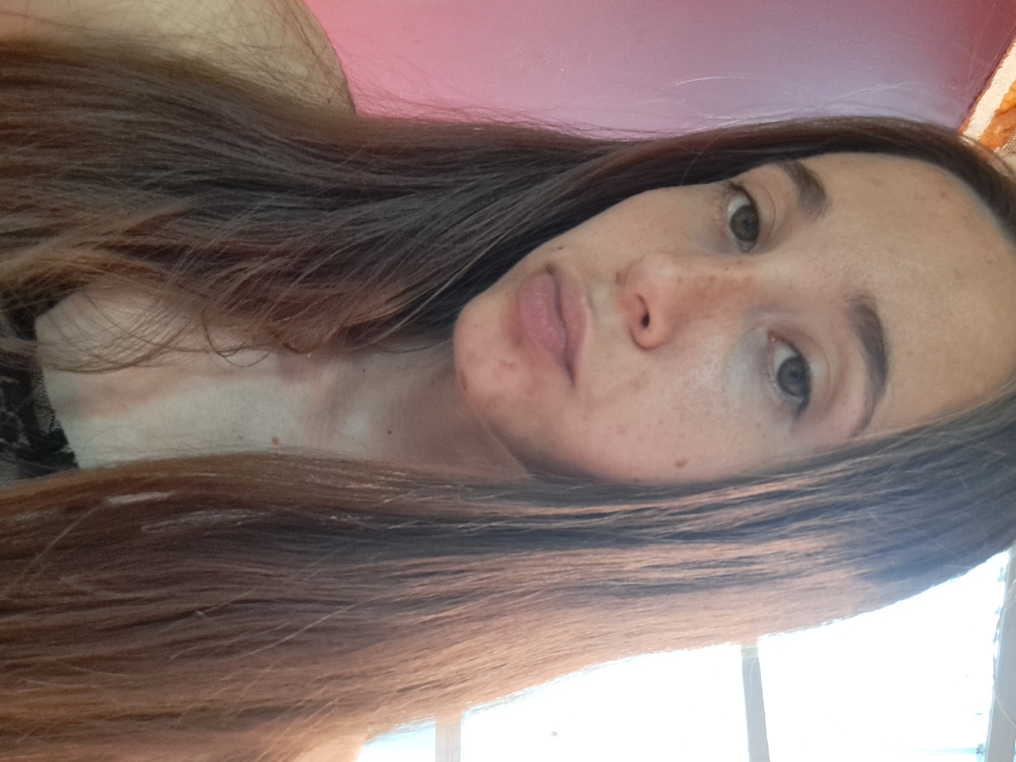 A close-up, head-and-shoulders selfie of a young woman with long, brown hair is shown. She is looking directly at the camera with a neutral expression. The background is a blurry pink wall on the left and a bright window on the right. The image appears to have been taken indoors, possibly in a residential setting. There are no discernible landmarks or features that would allow for the specific identification of a location within Vypasne, Ukraine.