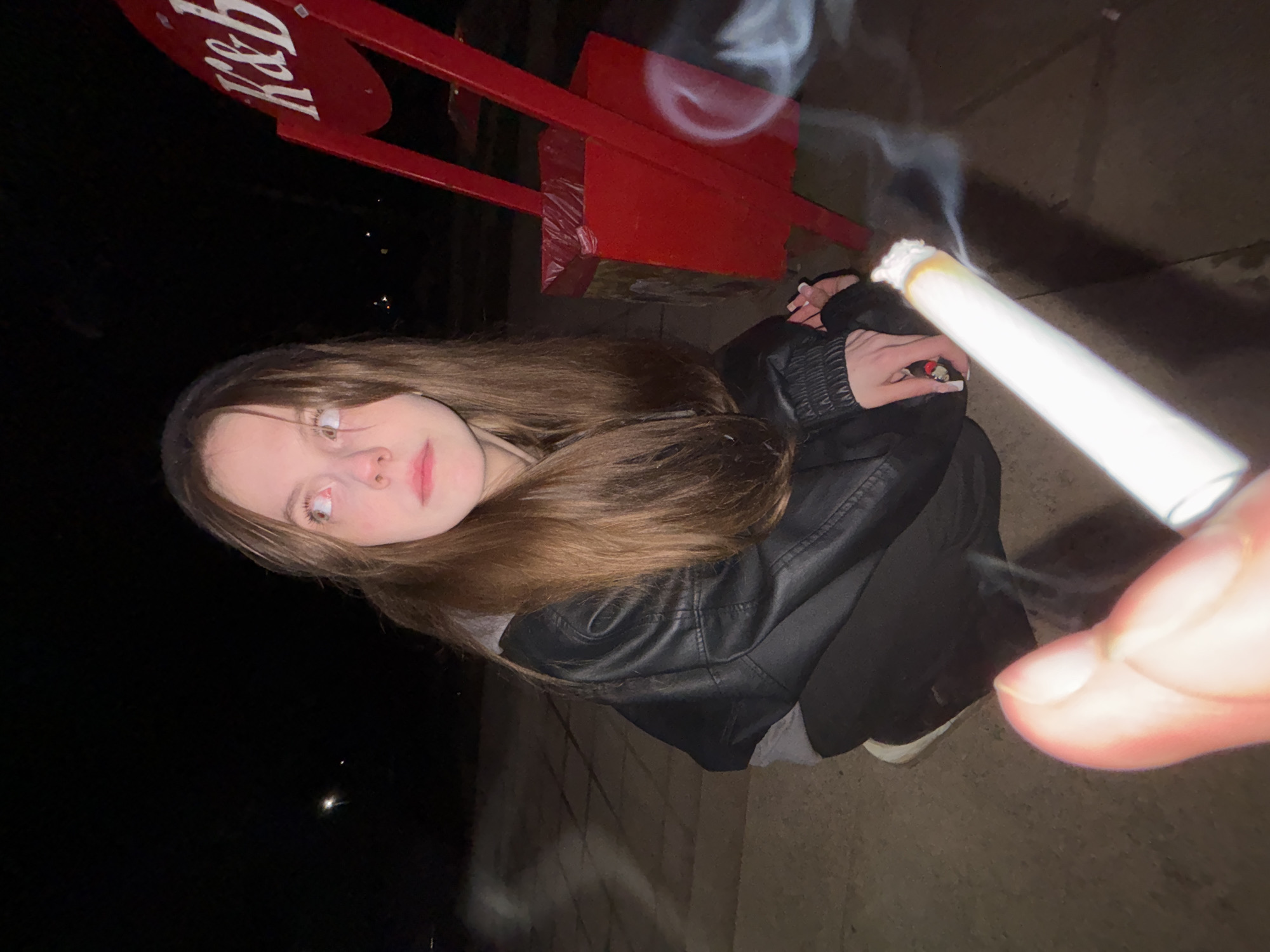 A young woman with long brown hair is crouched outdoors at night, holding a lit cigarette. She is wearing a black leather jacket and dark pants. In the background is a red sign with white letters, possibly related to a business or location. The location is not discernible in the city of Фоки, Russia. The environment appears to be a paved area or sidewalk.