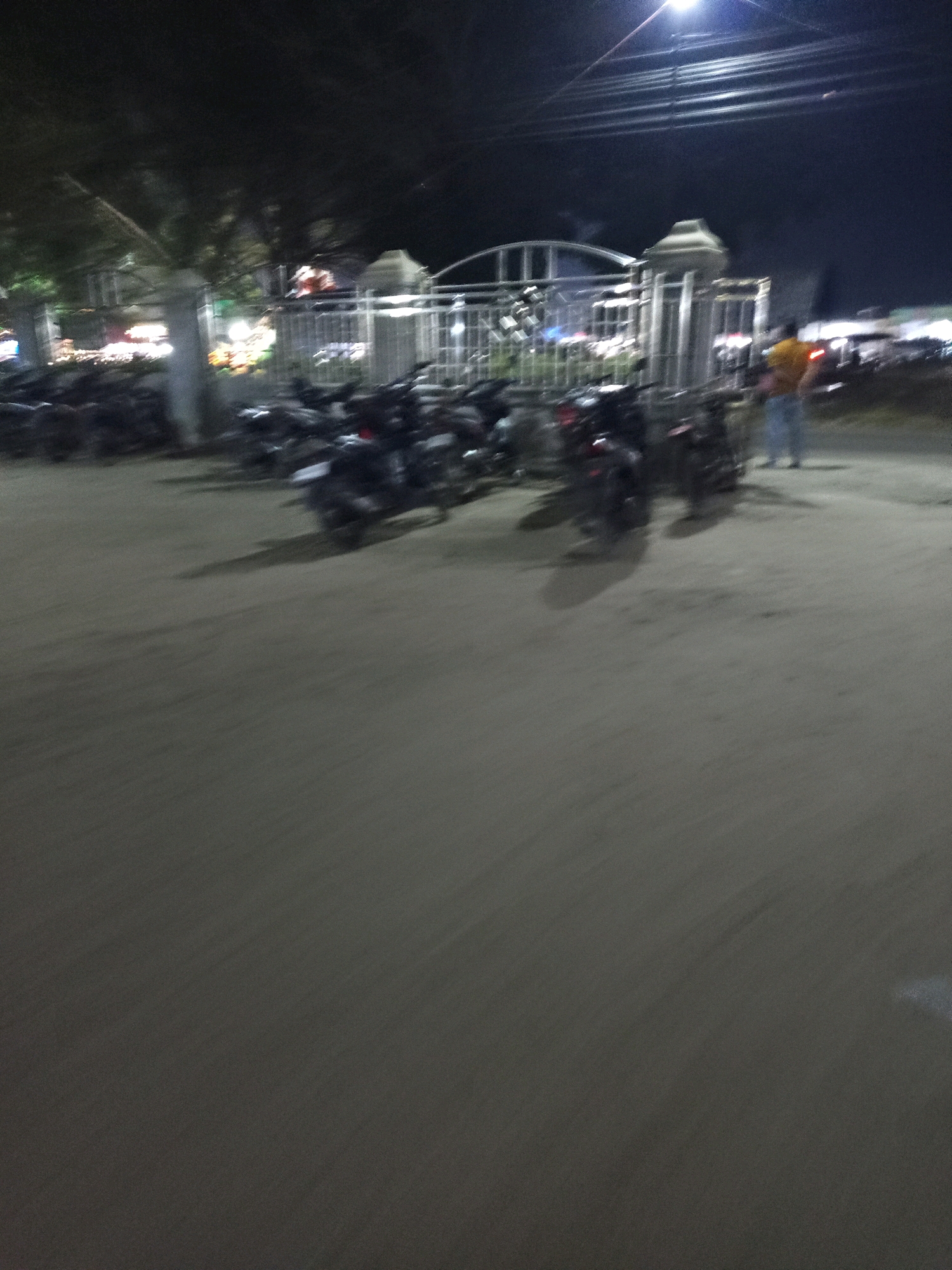 The image shows a dimly lit outdoor scene at night, likely in Pulung Kencana, Indonesia. A row of parked motorcycles is visible in the foreground, with their headlights and taillights casting faint glows. Behind the motorcycles, a metal gate with decorative posts stands open. A person wearing a yellow shirt and blue pants is walking away from the camera, towards the gate. In the background, trees are silhouetted against a dark sky, and some blurry lights suggest the presence of buildings or other activity. The ground appears to be paved, and shadows are cast by the motorcycles and the gate. The overall impression is one of a quiet, possibly public, area at night, with limited activity.