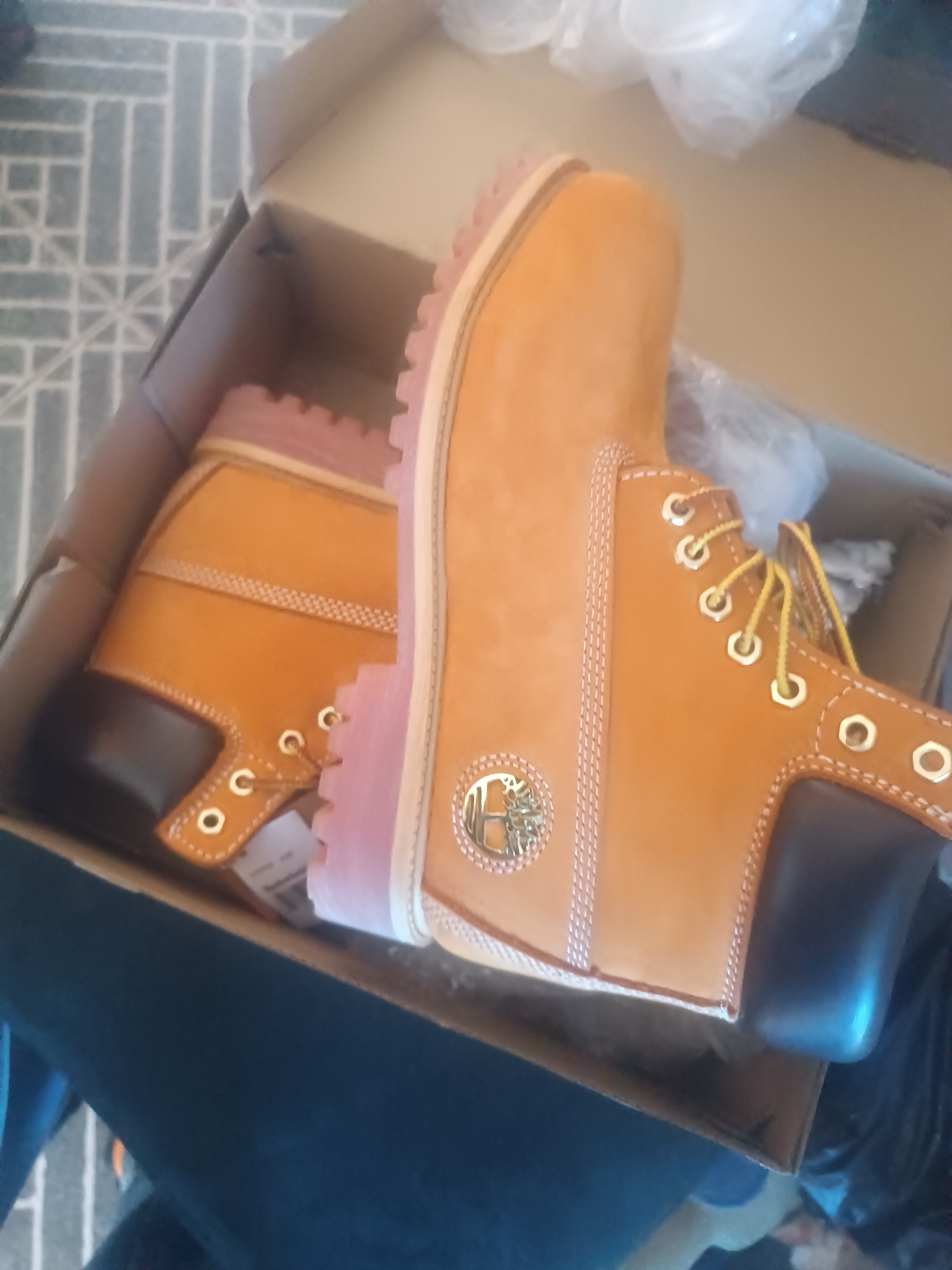 Two Timberland boots are in a shoebox. The boots are tan with pink soles, black heels and gold eyelets. They are sitting on a gray, geometric patterned floor. The box is in Bukuru, Nigeria.