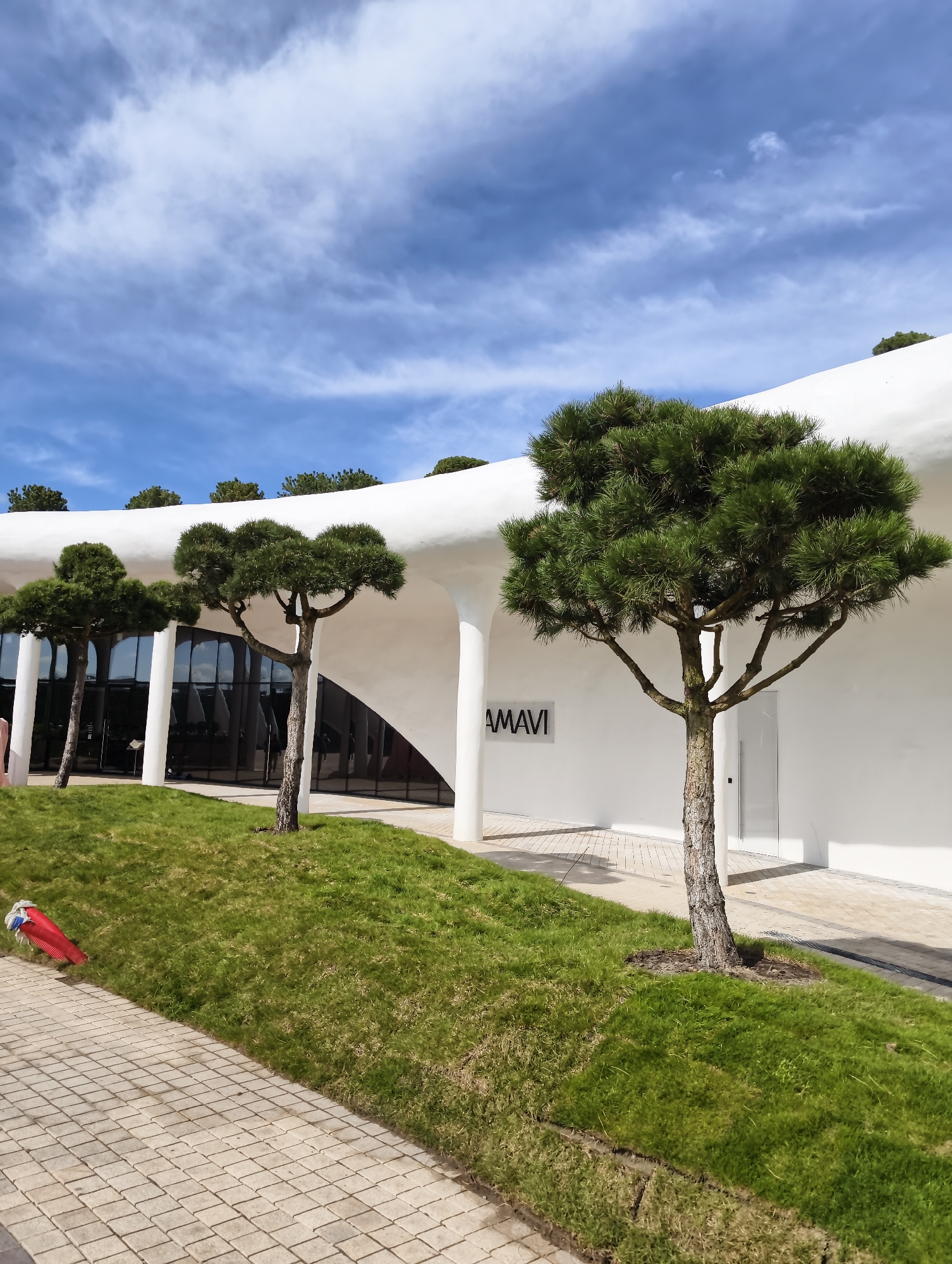 This vertical outdoor shot captures a modern, minimalist architectural scene under a bright, partly cloudy sky in Krasnodar, Russia. The central subject is a flowing, white building characterized by smooth, curved lines and archways, suggesting a contemporary design. Large, dark-tinted glass panels form the facade of the building, reflecting parts of the surroundings.
In the foreground and middle ground, several distinctively shaped, pine-like trees with dense, green foliage and textured trunks are strategically placed, enhancing the aesthetic of the landscape. A sloping, well-maintained green lawn occupies the area between the viewer and the building. Along the bottom left, a paved pathway made of light-colored, square-patterned blocks is visible. A small red object, possibly a hose or cable, is partly visible on the edge of the grass and pavement on the left.
The sky overhead is a clear blue with scattered, wispy white clouds. The bright daylight illuminates the scene, casting soft shadows, particularly from the trees and the building's overhangs. On the white facade of the building, between two columns, the word "AMAVI" is displayed in a dark, simple sans-serif font. The overall impression is one of modern design, meticulous landscaping, and a serene outdoor environment. No people or specific activities are visible.