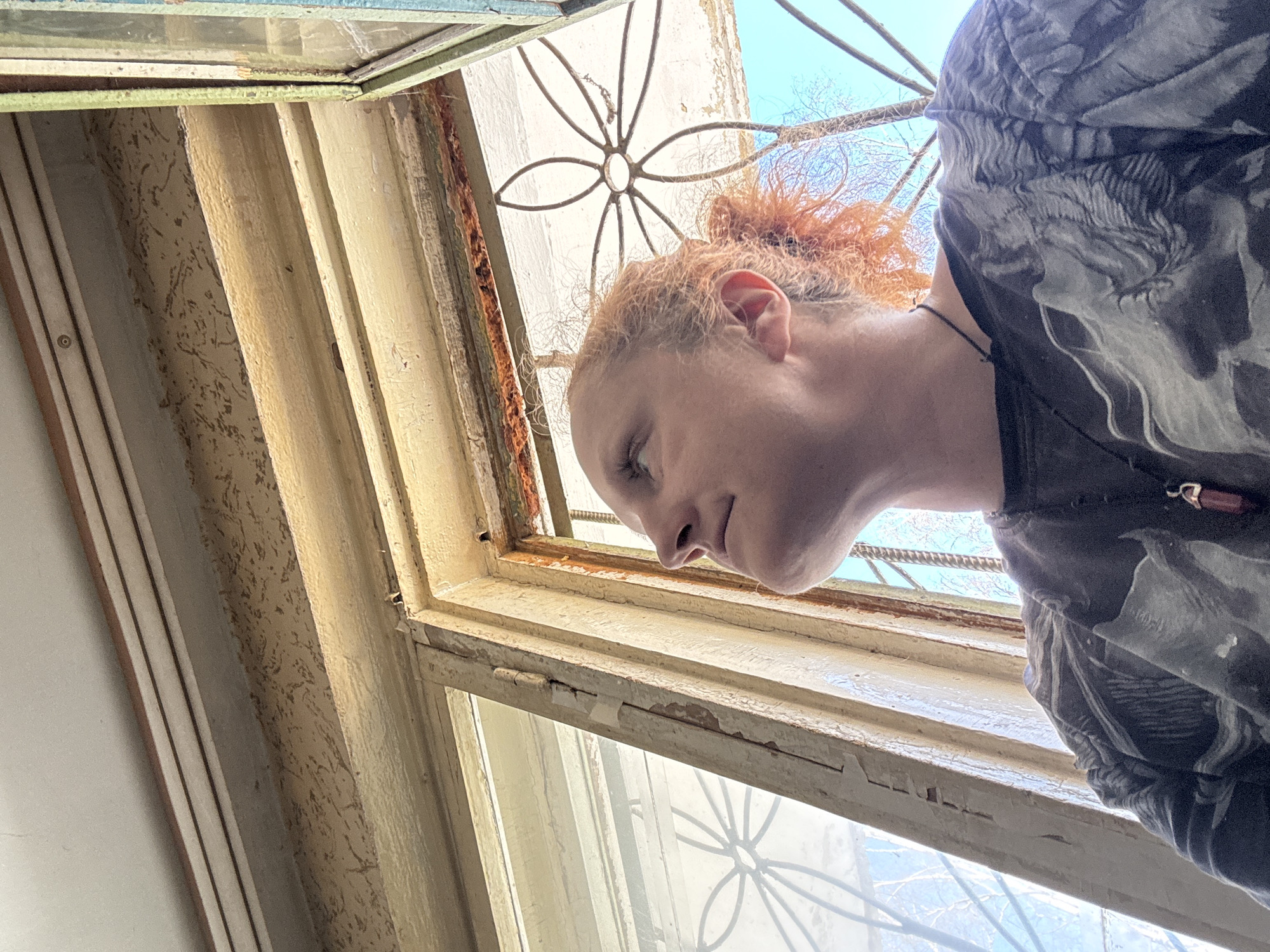 The image presents a low-angle view of an individual positioned in front of a window in Chaikovsky, Russia. The subject, a person with reddish-orange, curly hair styled into a bun, is shown in profile, looking upwards and to the left. They wear a dark t-shirt with a prominent light-colored, possibly abstract or animal-themed, graphic design across the upper torso. A black cord necklace with a reddish-brown, elongated pendant hangs around their neck.
The window frame is constructed of old wood, painted a faded, yellowish-beige color, with significant peeling and chipping paint that reveals darker underlayers. Ornate, dark-colored metal security bars, designed in a multi-petal floral pattern, are visible through the glass. Beyond the window, a clear blue sky is apparent, punctuated by the silhouettes of leafless tree branches. Above the window frame, a section of a light-colored interior ceiling or wall is visible.