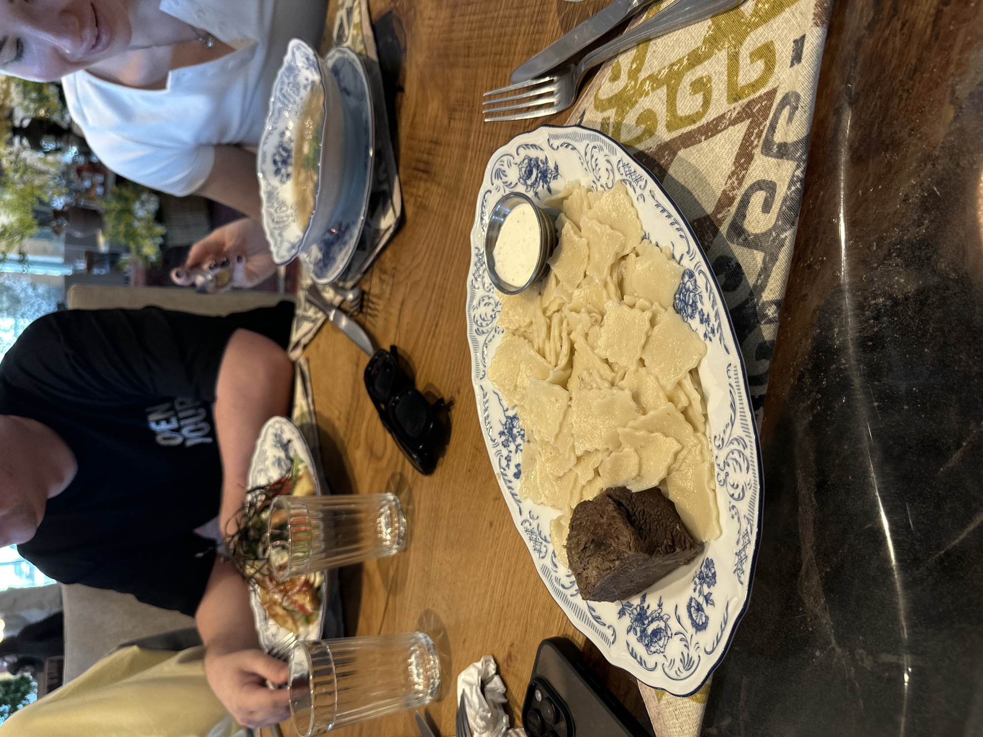 This image captures a casual dining scene, likely in a restaurant or cafe, with a focus on food served on a wooden table. The setting includes patterned fabric placemats with geometric designs in green, brown, and blue tones.
In the foreground, a large oval plate with distinctive blue and white floral patterns holds a traditional dish. It contains numerous small, irregularly shaped dough pieces, resembling a type of dumpling or pasta (possibly *khinkal*, a Dagestani specialty). Alongside the dough, there's a substantial piece of dark, cooked meat or blood sausage. A small metal cup filled with a creamy white sauce, likely sour cream or a similar condiment, is also on the plate.
Other dishes are partially visible: a bowl with blue patterns, possibly containing a soup or stew, and another plate holding a salad. Several ribbed glass tumblers are placed on the table, some appearing empty. Utensils, including forks and knives, are neatly arranged beside the plates.
Two individuals are partially visible at the table. One person on the upper left wears a white top, with only their lower face visible, suggesting they are looking down at their food. Another person on the lower left wears a black t-shirt, with part of their arm and shoulder visible. The text "DEN JOUR" is partially legible on the t-shirt. Personal items on the table include a black pair of sunglasses and a black smartphone (appears to be an iPhone).
The background is softly blurred, showing an indoor environment with hints of greenery, suggesting plants or decorations. The overall atmosphere appears relaxed, indicative of a shared meal. The food presented, particularly the dough pieces with meat and sauce, aligns with the culinary traditions found in Novo-Zubutli, Dagestan, Russia.