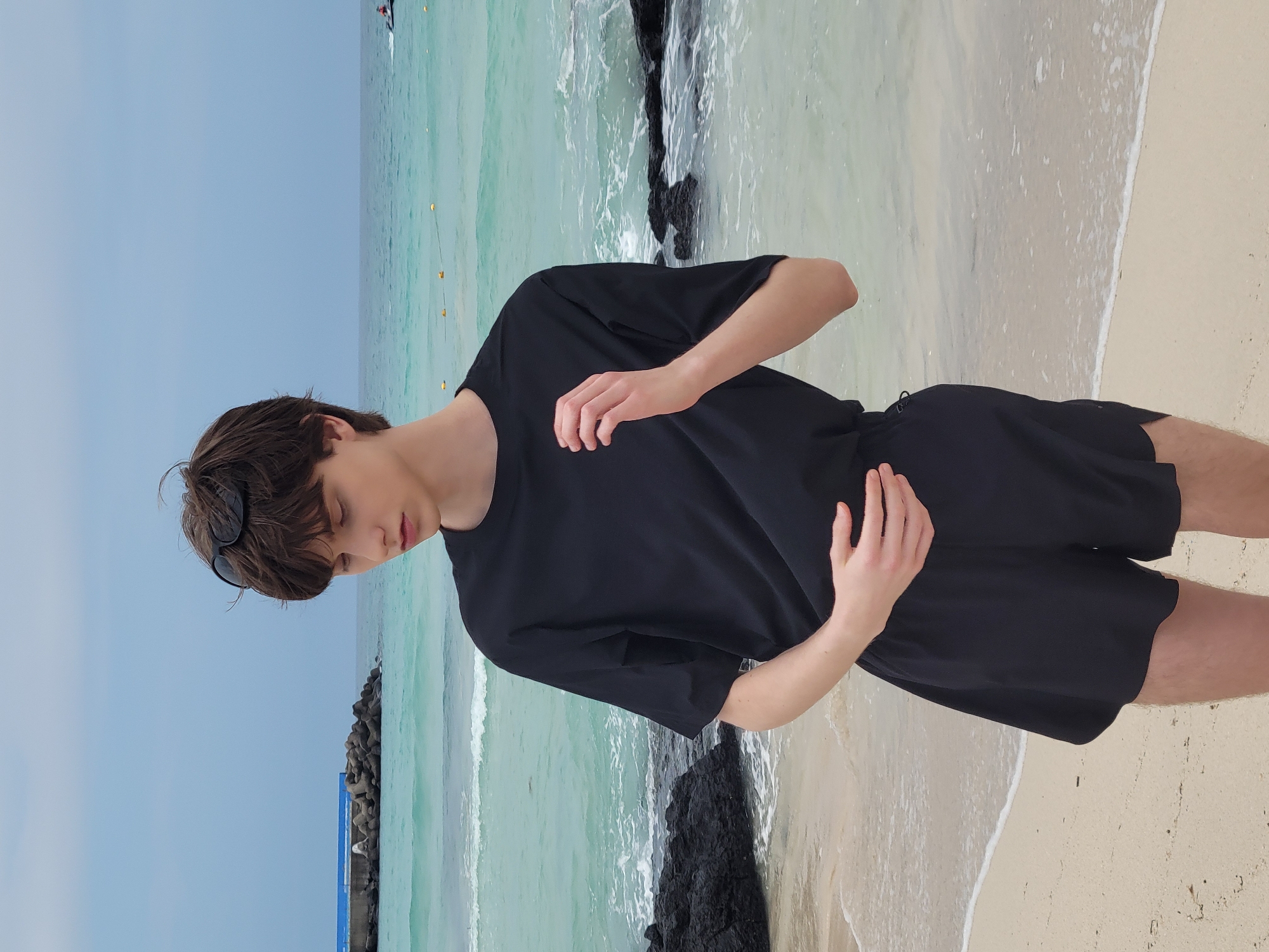 A young person with dark hair and fair skin stands on a sandy beach in Jeju, South Korea. They are wearing a black t-shirt and black shorts. Sunglasses are perched on top of their head. The person is looking down and has one hand on their stomach and the other near their chest. The ocean is a bright turquoise with white waves, and there are dark rocks on the shore. In the distance, there is a structure that appears to be a breakwater with a blue element on top, and a jet ski is visible on the water. The sky is overcast.