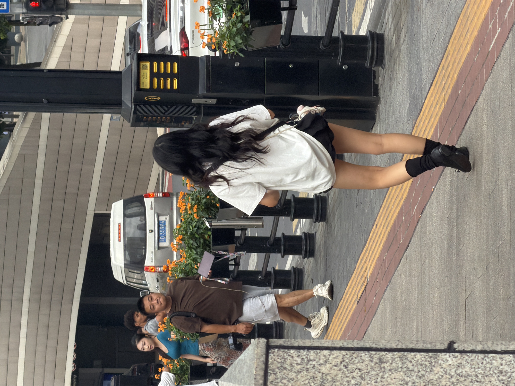 The image depicts a street scene in Yuzhong District, China, with several individuals and vehicles visible. In the foreground, a young woman with long, dark hair walks away from the camera, wearing a white t-shirt, black shorts, black socks, and black shoes. She carries a black bag over her shoulder. To the left of the frame, a man wearing a brown t-shirt, grey shorts, and white sneakers walks towards the camera, holding a purple rectangular object in his right hand. Behind him, a woman in a blue tank top and a patterned skirt walks towards the left. A white van with a license plate reading "JD 359P6" is parked in the background, along with a white car. The street is paved with grey concrete and features a yellow and red tactile paving strip along the edge of the sidewalk. Orange flowers in black planters line the walkway. A black lamppost with a control panel is situated to the right of the frame. The environment appears to be an urban setting with a building facade in the background featuring horizontal beige and grey stripes.