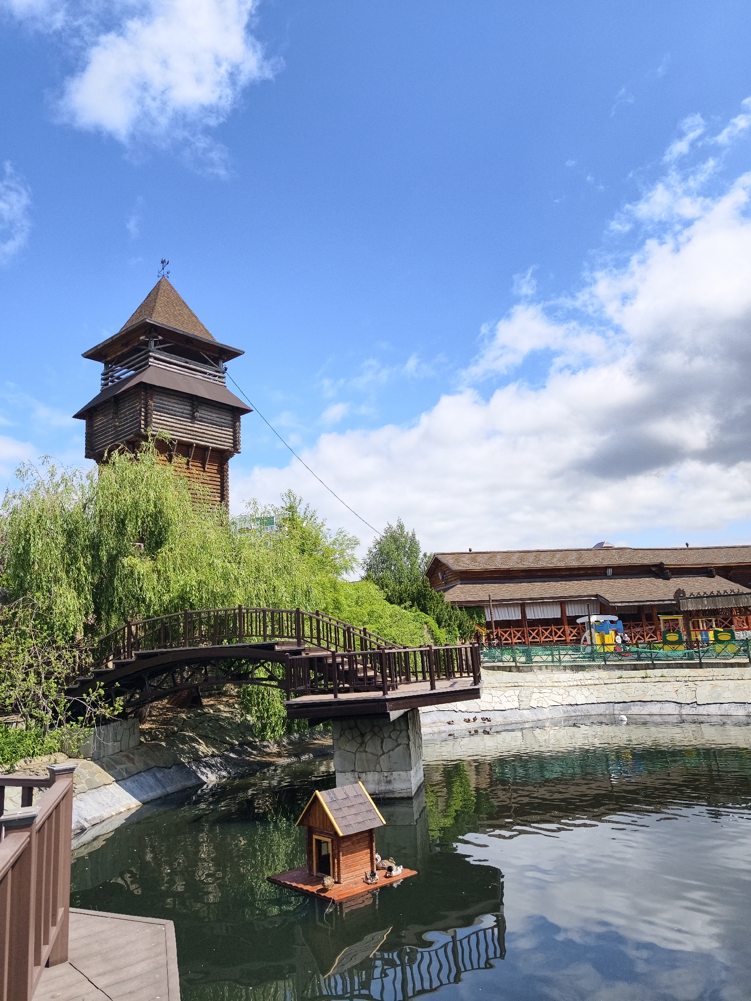 This image depicts a picturesque and tranquil outdoor scene, likely a recreational area or park in Izhevsk, Russia, under a bright blue sky with scattered white clouds. The primary subject is a tall, multi-tiered wooden observation tower with a brown shingle roof, crowned by a weathervane. Abundant green foliage, including large willow trees, surrounds the base of the tower and lines the water's edge.
A gracefully arched wooden footbridge with dark railings spans a section of a pond or small lake. In the foreground, a small, charming wooden house designed for ducks floats on the water, with a few ducks resting on its platform. More ducks are visible swimming in the pond and standing on the far bank.
Across the water, a long, multi-gabled wooden building with a similar brown roof suggests a restaurant or visitor facility. It features an open-air veranda with decorative wooden cross-patterned railings and colorful play equipment or decorations on the paved area in front. The water reflects the sky and surrounding structures, enhancing the serene atmosphere.
The scene is well-lit by natural daylight, indicating a clear, pleasant day. There is no discernible human activity or visible text in the image.