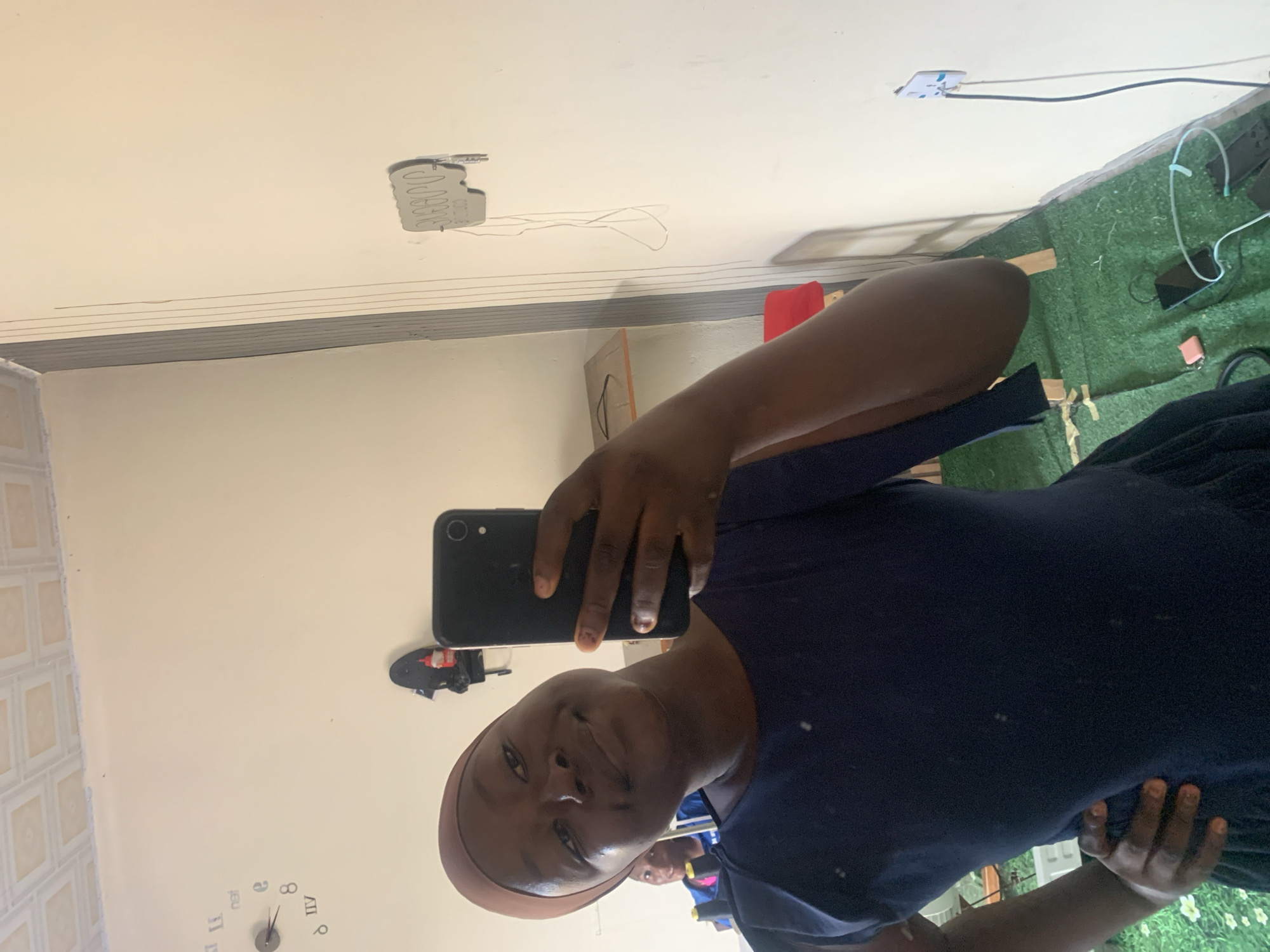 A medium shot captured in Jauro Yinu, Nigeria, shows a young Black woman taking a selfie in a room. She is wearing a dark blue dress and a light brown head covering. Her left hand is on her hip, and she is holding a black smartphone in her right hand, angled to capture her reflection in a mirror. Her expression is a subtle smile. In the background, another young Black woman is visible in the mirror, wearing a blue and pink top, looking towards the camera. The room has light-colored walls and a tiled ceiling. A wall clock is visible on the left wall, along with some shelves holding items including what appears to be a skateboard with a bottle on it. A wooden shelf unit with a red object on it is also visible. The floor is covered with artificial grass. Electrical outlets and cords are present on the right wall.