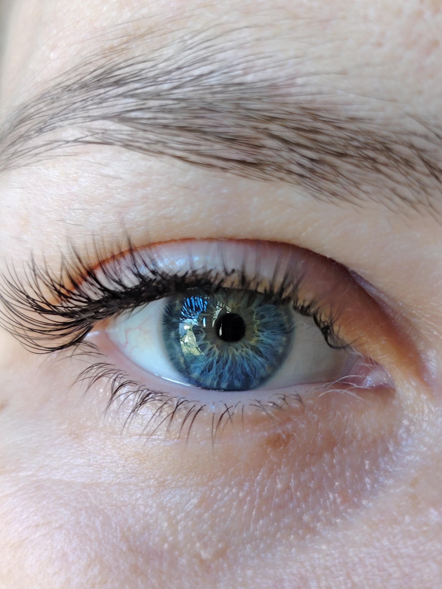The image is a close-up of a human eye. The eye is blue with a black pupil. The iris shows intricate patterns. The eyelashes are dark and prominent. Above the eye is an eyebrow with individual hairs visible. The skin around the eye is light and shows texture and fine wrinkles. There is no information available to suggest the location in the city of Chaikovsky, Russia.