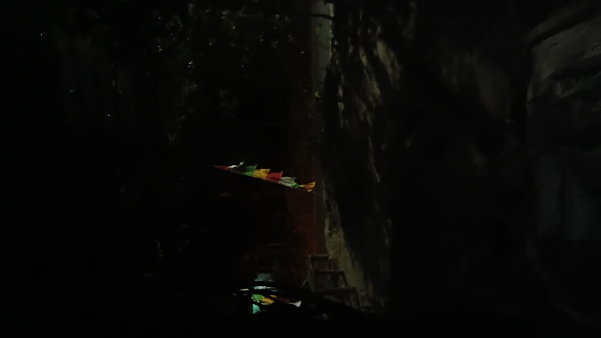The image depicts a dark outdoor scene, likely at night, in Prabumulih, Indonesia. A string of colorful pennant flags, red, yellow, green, and white, hangs from a pole on the right side of the frame, swaying slightly. To the left, a dark silhouette of a structure with a table or bench in front of it is visible, partially obscured by shadows. The ground appears to be sandy or dusty, with scattered debris and patches of light and shadow cast by an unseen light source. Trees and foliage are visible in the background, also largely in shadow. The overall impression is one of a dimly lit outdoor area, possibly a park, roadside, or communal space.