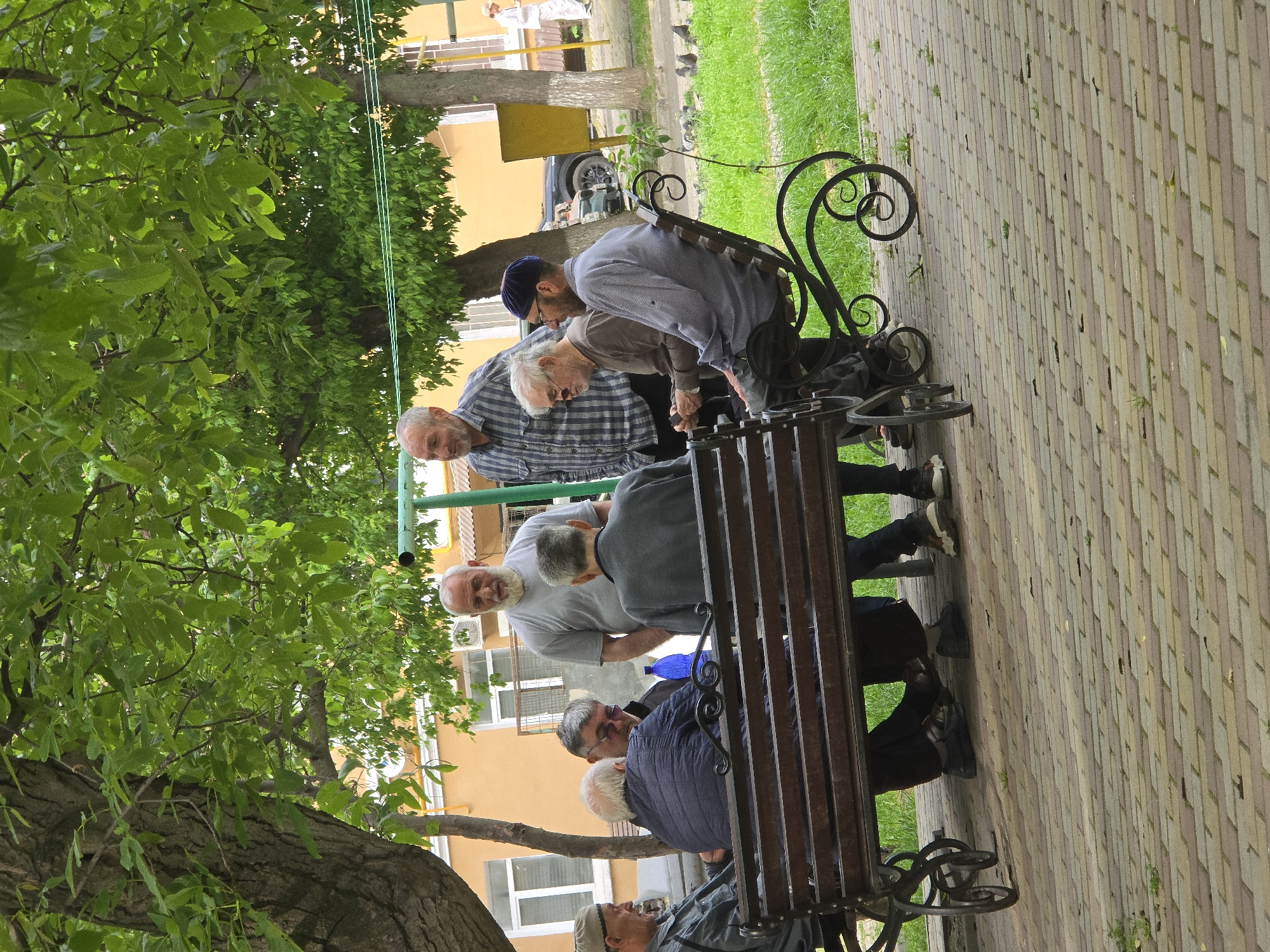 A group of elderly men are gathered around a park bench in what appears to be the city of Grozny, Russia. The bench is made of dark wood and ornate metal, with a curved back and armrests. The men are dressed in casual clothing, including shirts, pants, and jackets. Some of them are wearing hats or caps. They are interacting with each other, some leaning over the bench and others sitting. A tree with green leaves provides shade over the scene. The ground is paved with brick. The location is likely in Grozny, Russia, given the cultural attire of the men and the architecture in the background. A residential building with white-framed windows is visible behind the bench.