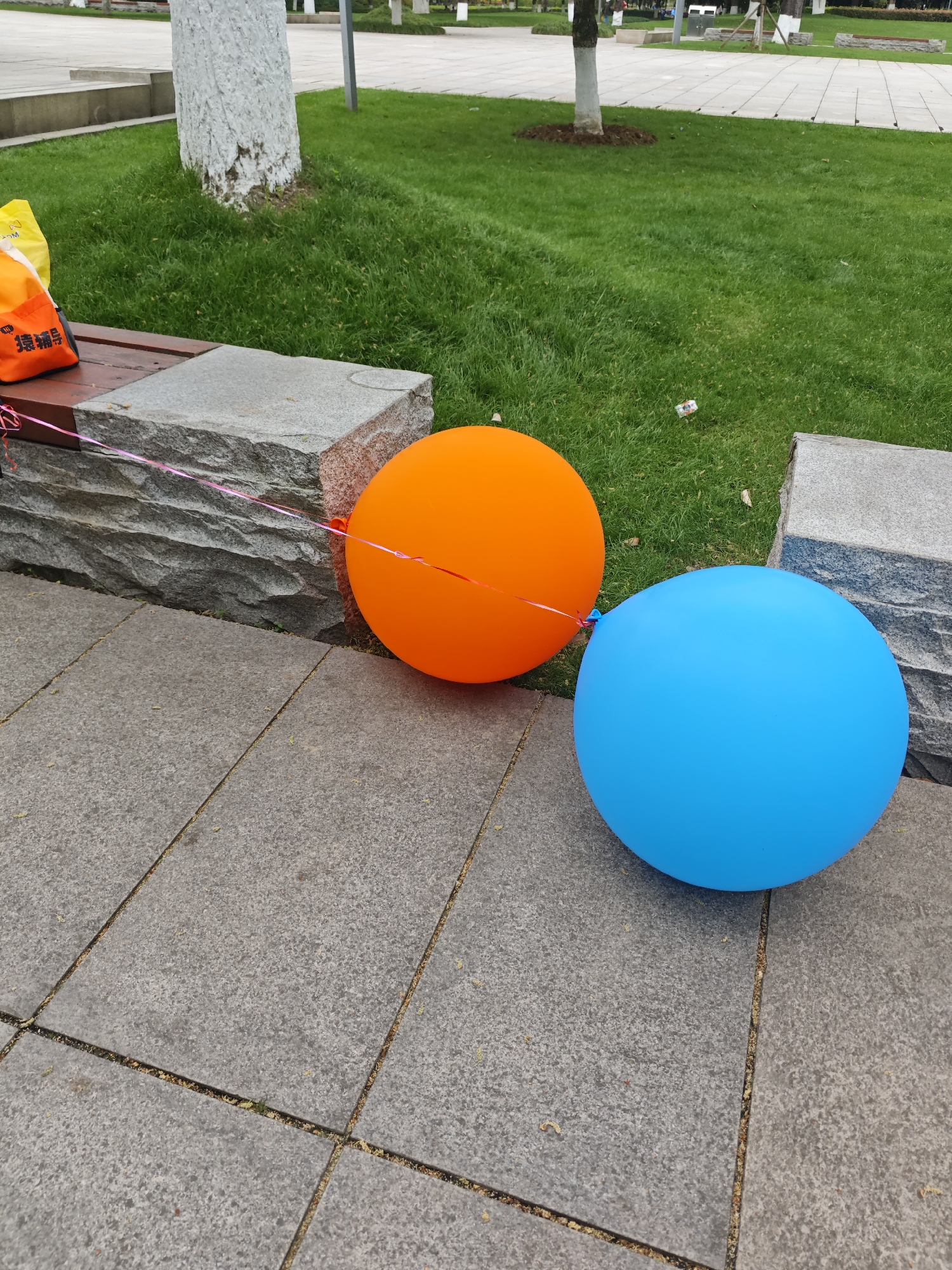 The image shows an outdoor scene in Liangjiang, China, featuring two balloons, one orange and one blue, resting on a gray tiled surface. The balloons are situated near a low stone bench with a package resting on top. Behind the bench is a patch of green grass with trees in the background. The lighting suggests it is daytime. No people are visible in the frame.