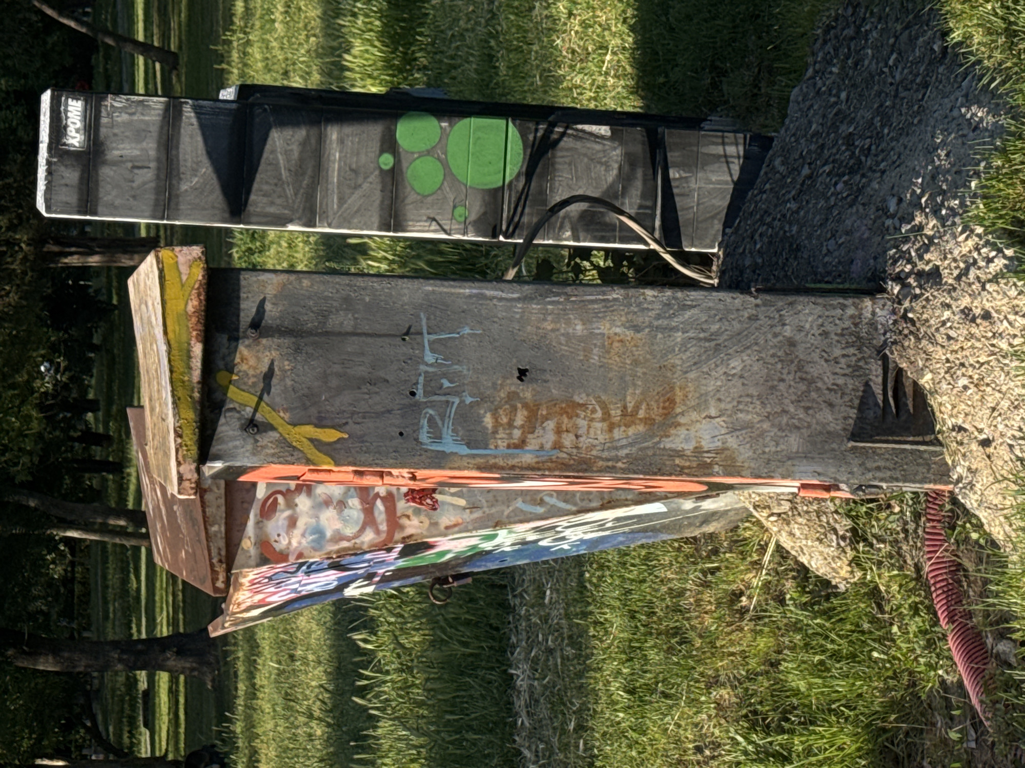 The image shows a heavily vandalized metal utility box sitting on the ground amidst grass and debris. The box has multiple layers of graffiti in various colors. On top of it sits a black "XPOME" branded object with green circular markings. The surrounding environment is grassy with scattered rocks and small branches. A red corrugated tube is also visible near the right side of the box. The location is likely a park or green space, potentially in Sofia, Bulgaria. The box is weathered, suggesting it has been in place for a considerable time. There are no people visible in the image.