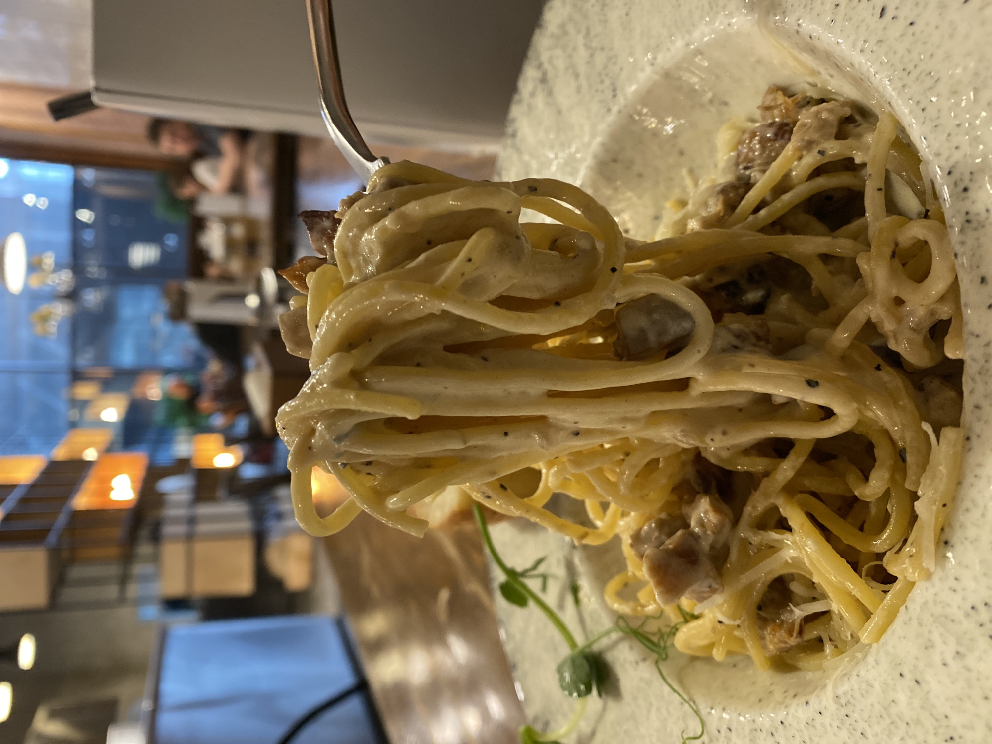 The image shows a plate of spaghetti carbonara held up by a fork. The pasta is coated in a creamy sauce with visible black pepper and pieces of bacon or ham. Some green sprouts garnish the plate. The plate itself is off-white with dark speckles.
In the blurred background, a restaurant setting is visible. Tables and chairs are arranged, and there is a window reflecting a cityscape. The location is implied to be in the city of Yekaterinburg, Russia, based on the prompt. A person is partially visible in the background, possibly another patron.