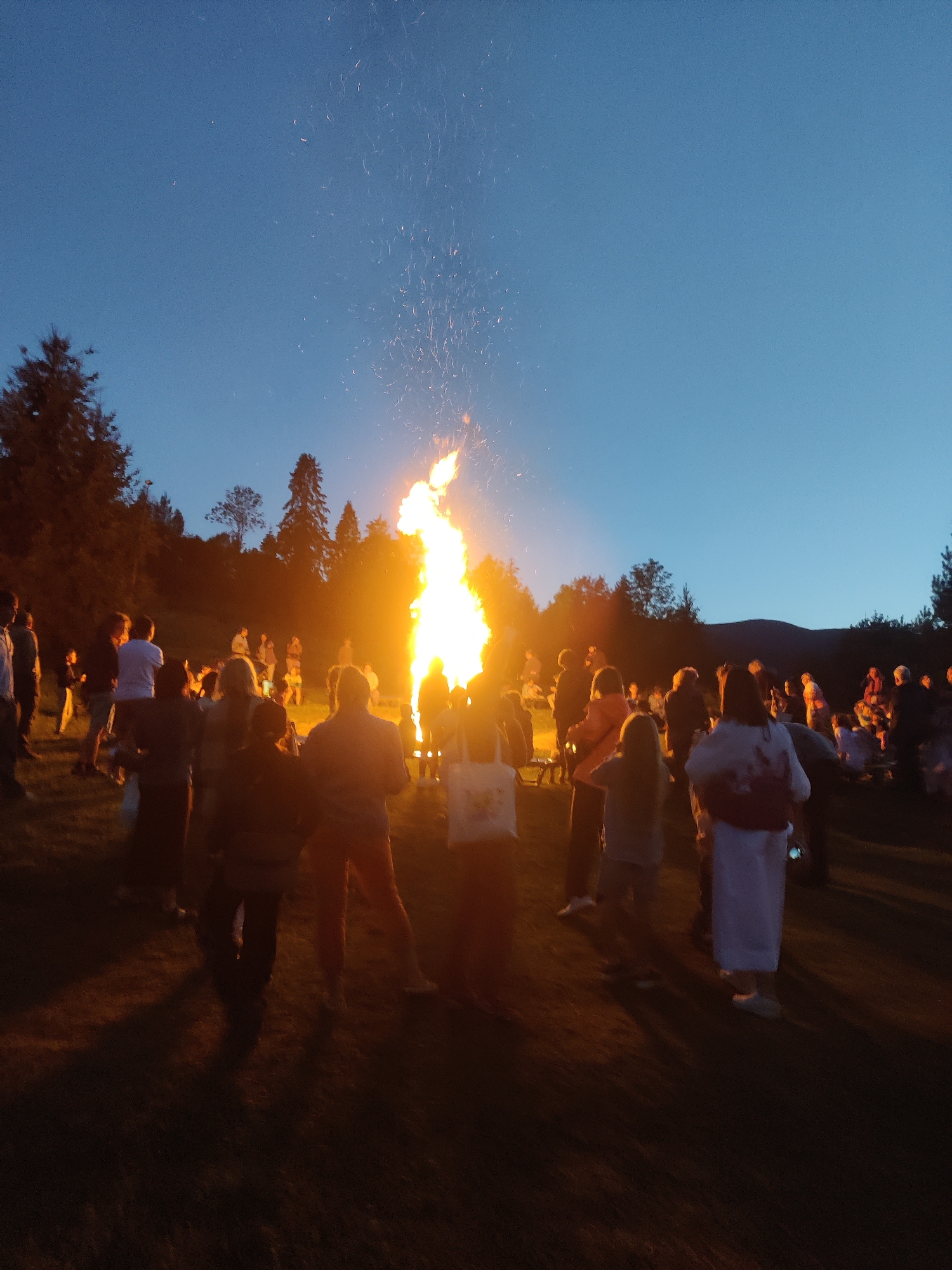 A group of people are gathered around a large bonfire at dusk in Izky, Ukraine. The bonfire is the central focus of the image, with flames reaching high into the dark blue sky, and sparks flying upwards. The surrounding area is dimly lit by the fire, with silhouettes of trees visible on a hillside in the background. The people are standing or sitting on the grass, most of them facing the bonfire. Some individuals are talking or looking at the fire, while others appear to be observing the spectacle. The overall atmosphere seems to be one of community and shared experience.