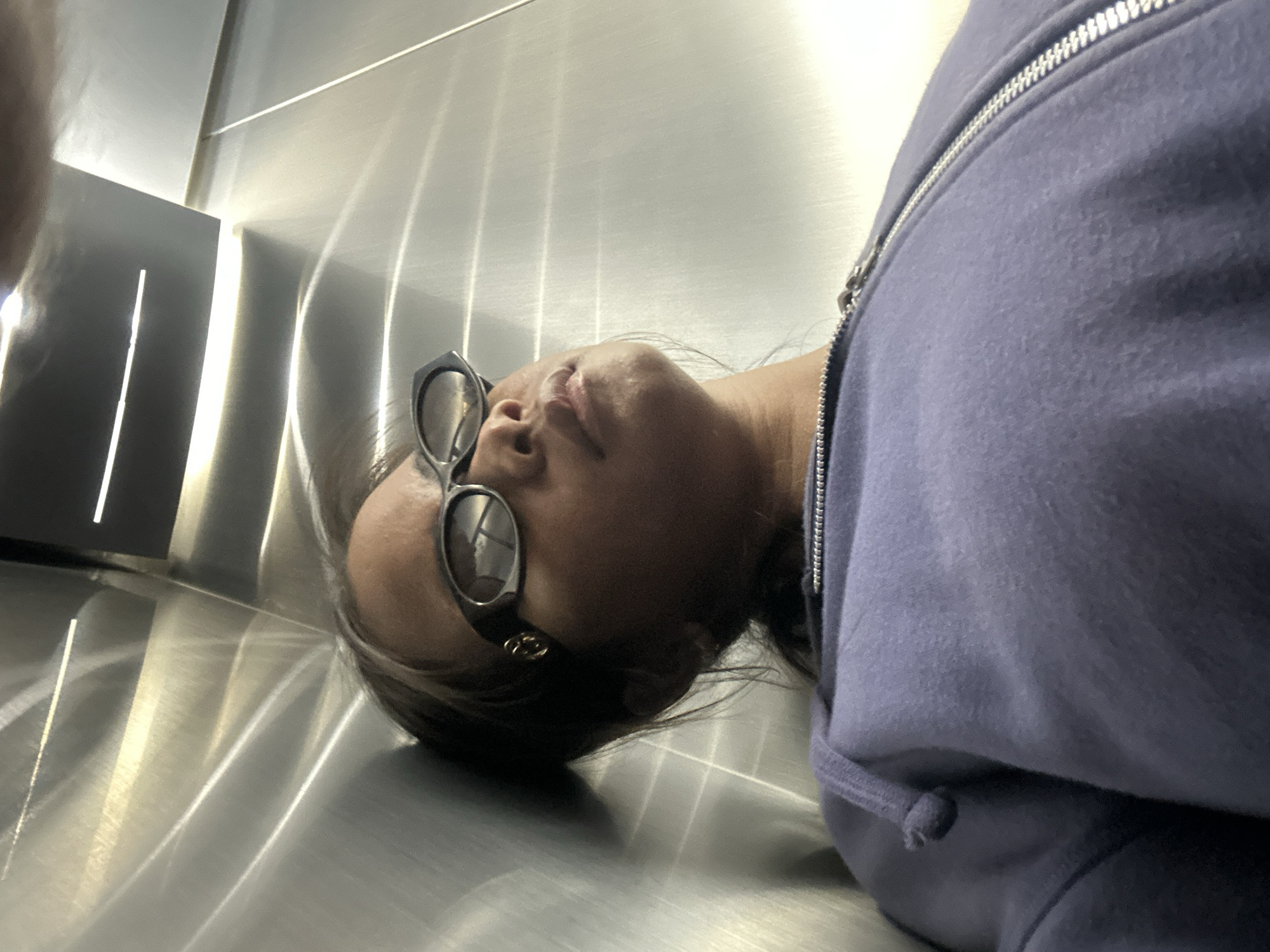 A person is leaning against a metallic wall in what appears to be an elevator. They are wearing sunglasses and a purple hoodie. The environment is reflective and shows streaks and patterns from the metallic surface. The lighting is artificial and casts shadows. The overall impression is that of a candid, possibly indoor shot in an urban setting. The question about Kuta, Indonesia is not supported by any visual evidence in the image.