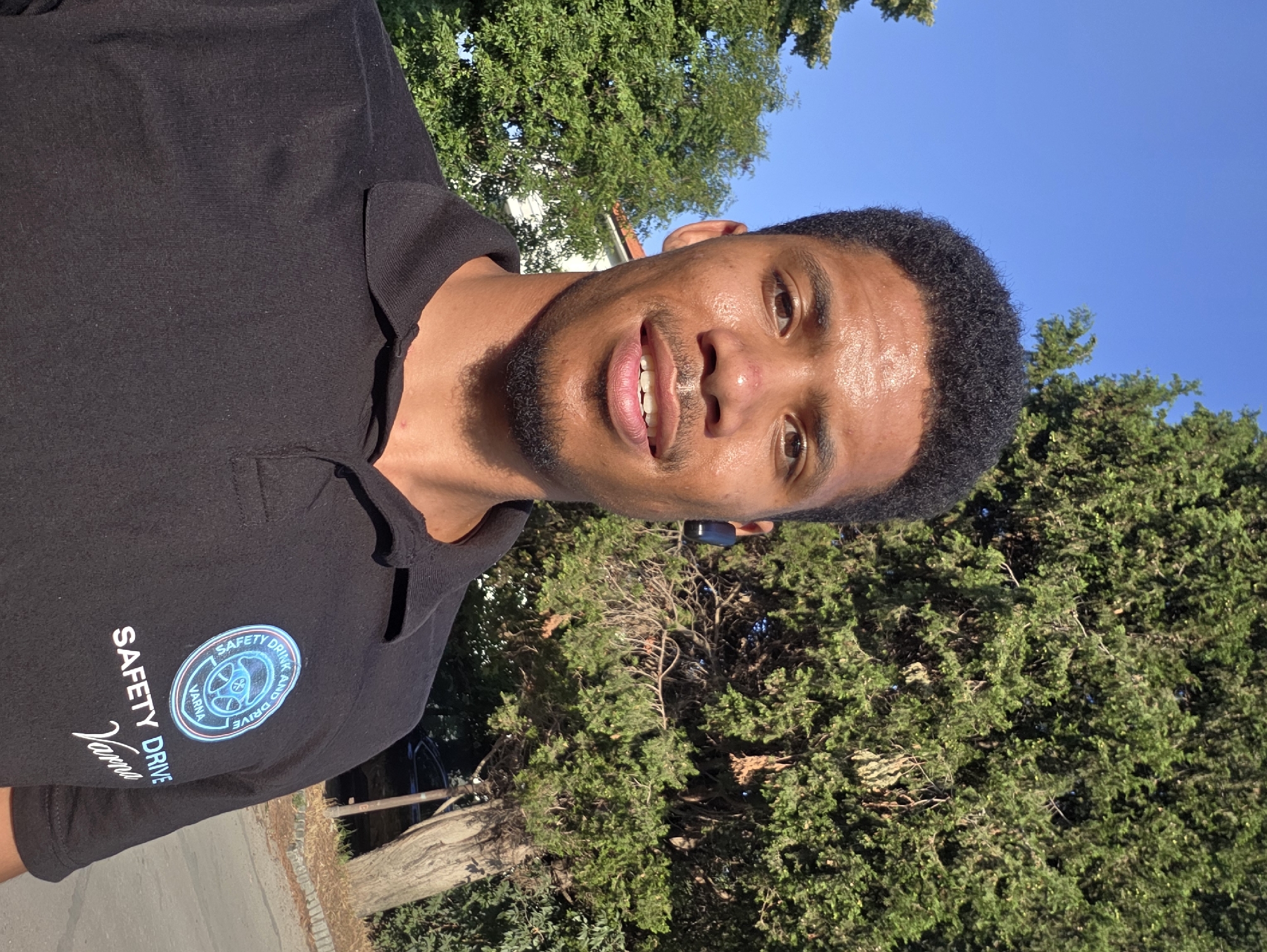 A young Black man is captured in a selfie outdoors with a clear blue sky and lush green trees in the background. He is wearing a black polo shirt with a circular emblem on the left chest that reads "SAFETY DRINK AND DRIVE VARNA." Below the emblem, it says "SAFETY DRIVE VARNA" in white lettering. He has short, dark, curly hair and is wearing a black wireless earbud in his left ear. His head is turned slightly towards the camera, and he has a light beard and a slight smile. The sunlight illuminates his face, and his skin appears slightly sweaty, suggesting warm weather. The backdrop consists of dense green foliage of trees, with a glimpse of a building and a parked car visible in the distance to the left and right, respectively. The overall environment suggests a park or street in Varna, Bulgaria, on a sunny day.