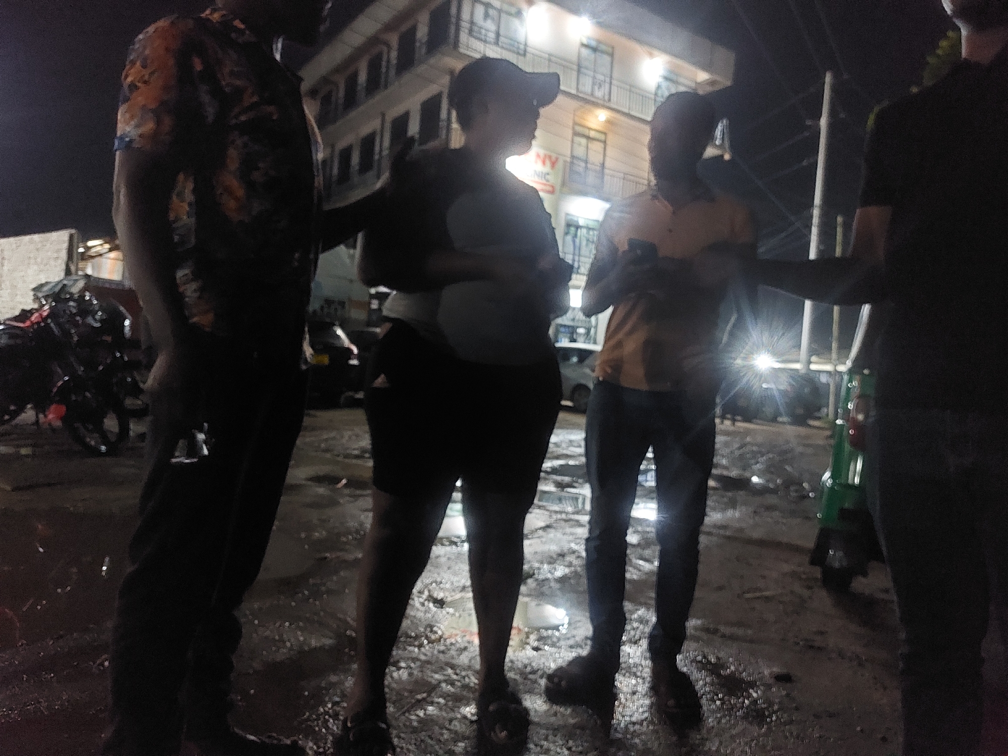 The image depicts a group of four people standing on a wet street at night. The first person, on the left, is wearing a patterned shirt and dark pants. The second person is wearing a baseball cap, a light-colored shirt, and dark shorts. The third person is wearing an orange shirt and jeans and is looking at a cell phone. The fourth person is on the right and is wearing a dark shirt and dark pants. In the background, there is a multi-story building with lit windows and a sign that reads "NY CLINIC". Motorbikes are parked in the street to the left. A green three-wheeled vehicle is parked on the right. The wet street suggests it has recently rained. The location is possibly in Dar es-Salaam, Tanzania, given the presence of the three-wheeled vehicle, which is common in that city.
