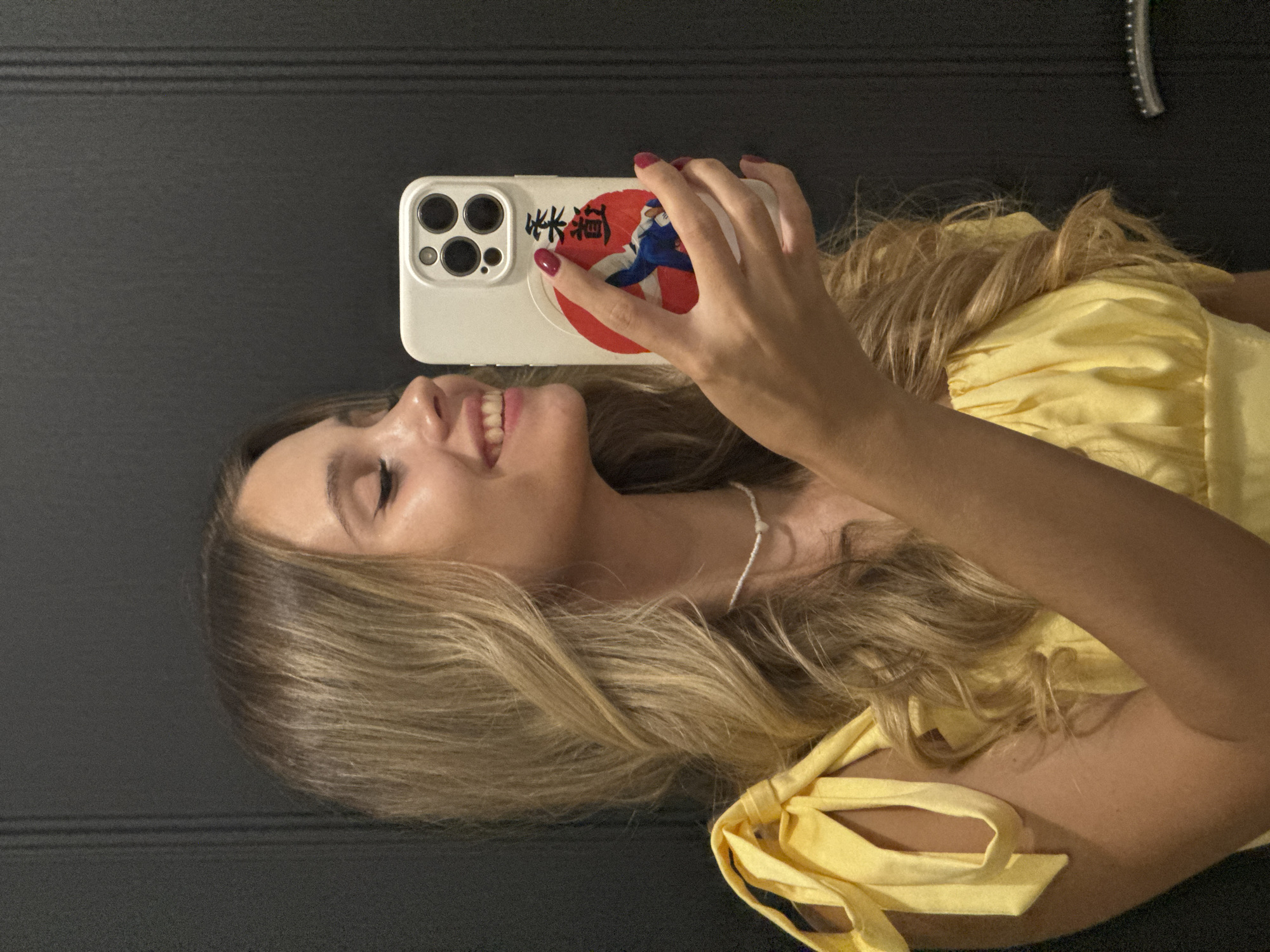 A young Caucasian woman with long, wavy blonde hair takes a selfie in a room with dark, vertical paneling on the right side, possibly a closet door or wall. She is wearing a yellow off-the-shoulder top with tie details on the straps and a white beaded necklace. Her left hand holds up a white phone with a camera lens array and a circular sticker featuring a person in a blue gi on a red background, surrounded by Japanese characters. Her right hand is extended, holding the phone in front of her. She is smiling and looking at the phone screen, with makeup visible on her eyes and lips, and red nail polish on her fingers. The image appears to be taken indoors, likely in a residential setting. There is no visible indication of the specific location in Chaikovsky, Russia, beyond it being an indoor environment.