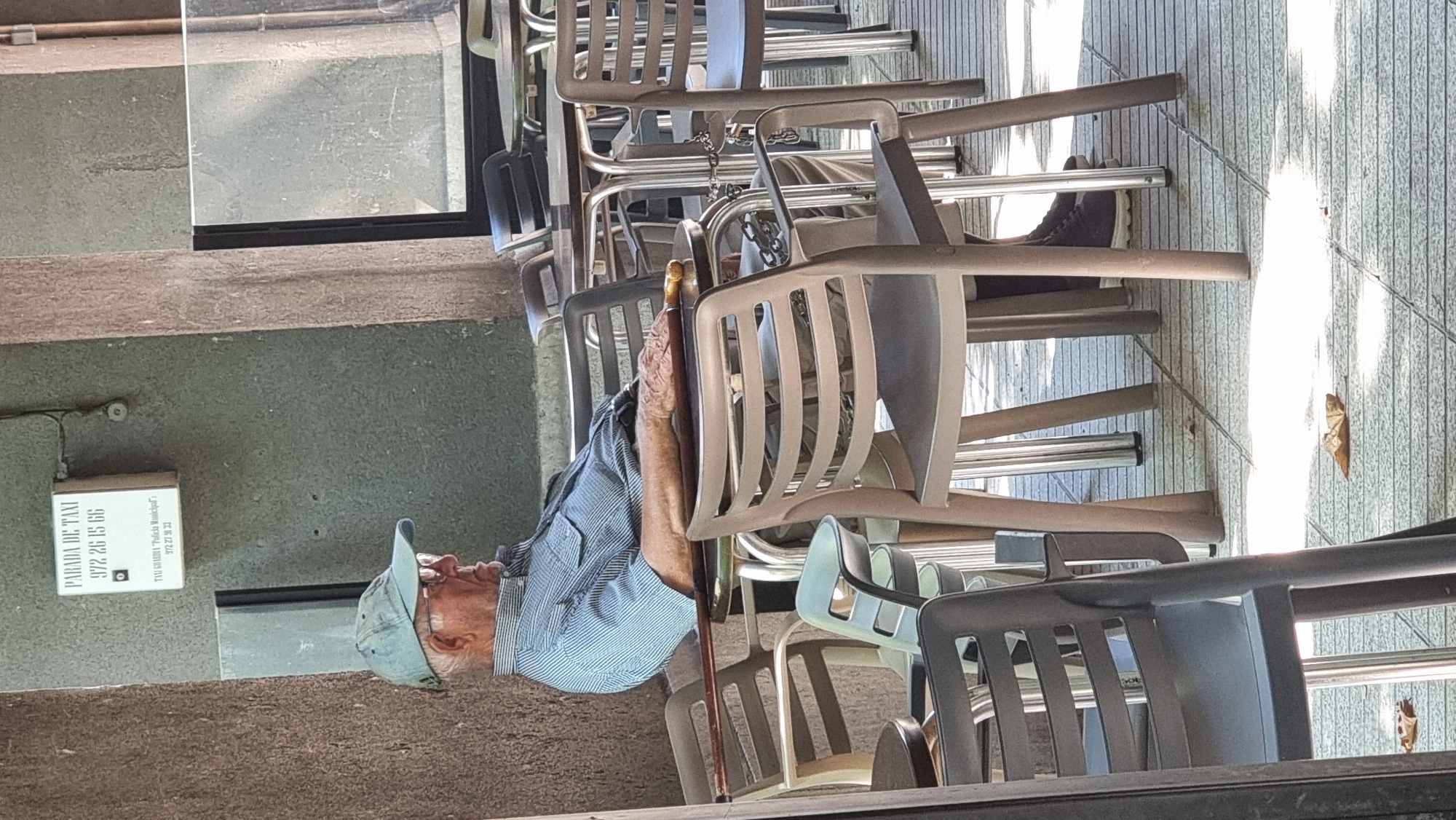 The image captures an elderly man sitting at an outdoor cafe table in Olot, Spain. He is wearing a light blue and white striped short-sleeved shirt, a light blue baseball cap, and glasses. His right arm is resting on a cane with a golden handle, which is placed across the table. The man appears to be looking to his right, with his eyes closed or looking down. The cafe setting includes multiple tables and chairs, some of which are empty. A sign for a taxi stand is visible in the background. The ground is paved with a tiled surface, and a few fallen leaves are scattered on the ground, suggesting an autumn or late summer setting. The scene is illuminated by natural sunlight, casting shadows across the pavement and furniture.