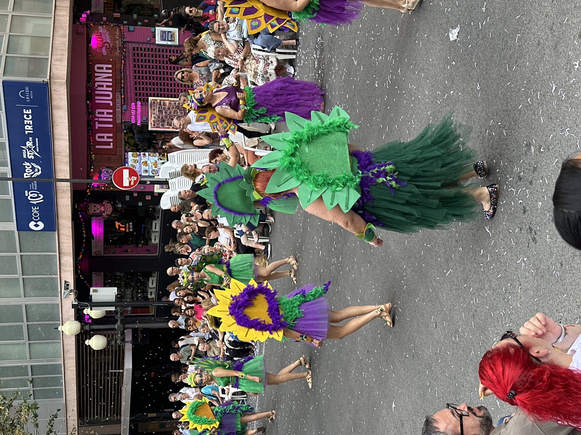 This image depicts a daytime street scene in an urban environment, likely a parade or carnival, set on an asphalt road scattered with small white pieces of paper or confetti. The location is **Spain, Unknown City**, as indicated by the prominent Spanish language on signs and the logos of Spanish media companies. In the foreground and midground, a group of approximately five to six female performers are walking from left to right. They are dressed in elaborate, colorful costumes predominantly featuring green, purple, and yellow. These costumes include short, layered tulle skirts (some green, some purple), bodices, and significant stylized headpieces or back decorations. Several performers wear large, yellow and purple elements resembling sunbursts or sunflowers, often accented with feathers. Other performers feature large, angular green shapes, possibly stylized leaves, also with green feathered boa accents. One performer in the center wears a purple skirt, a green feathered waistline, and a yellow-and-purple sunburst back piece, paired with golden flat sandals. Another performer in the foreground wears a green tulle skirt, a green feathered waistline, and large green angular back pieces, with black flats adorned with colorful embellishments. Along both sides of the street, numerous spectators are gathered, observing the procession. Many are seated on white plastic chairs, while others stand. The spectators are diverse in age and casually dressed. On the left sidewalk, a stack of unoccupied white plastic chairs is visible. In the extreme lower-left corner, parts of two spectators are visible: a man with a beard and glasses looking towards the right, and a woman with bright red hair looking towards the right. The background consists of multi-story urban buildings lining the street. On the right side, a ground-floor commercial establishment named "LA TIA JUANA," identified as "COMIDA MEXICANA," features a pink facade with neon lighting and a closed corrugated metal security shutter. Additional signage for "SOL Cerveza" and "OTRA VEZ ESTA MALDITA FELICIDAD" is present. A red circular "no entry" road sign is affixed to a pole. On the left side, another ground-floor business with horizontal slatted windows is partially visible, with a small sign indicating "NAKA CLUB." Small white, spherical objects, possibly bubbles, are suspended in the air in front of this building. Above the ground-floor businesses, a large blue sign displays logos for "COPE 100," "Rock FM," "MEGASTAR FM," "13CE," and "ABSIDE MEDIA." The upper floors of the buildings feature rectangular windows. Street furniture includes