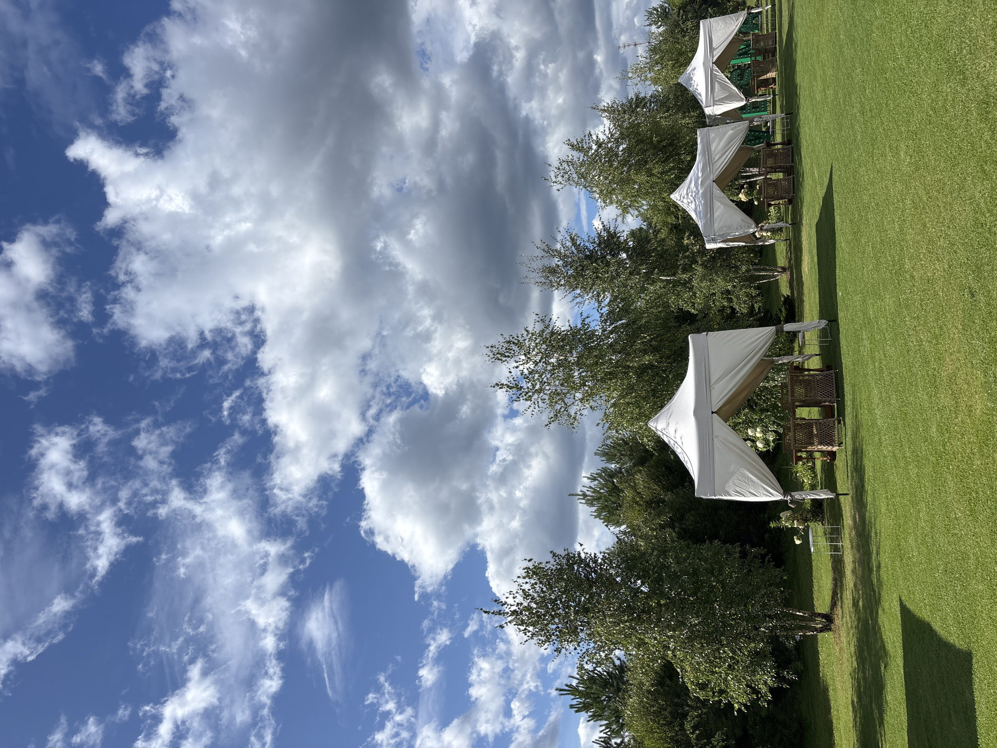 The image depicts an outdoor event setup on a bright, partly cloudy day in Pokrovskoe, Russia. The scene is a lush green lawn dotted with several white, peaked tents. Each tent appears to be an open-air pavilion, furnished with small tables and chairs, suggesting an outdoor dining or seating area. In the background, a dense line of various trees, including birches and evergreens, forms a natural backdrop. The sky is a vibrant blue, streaked with dramatic white and gray clouds. No people are visible in the image.