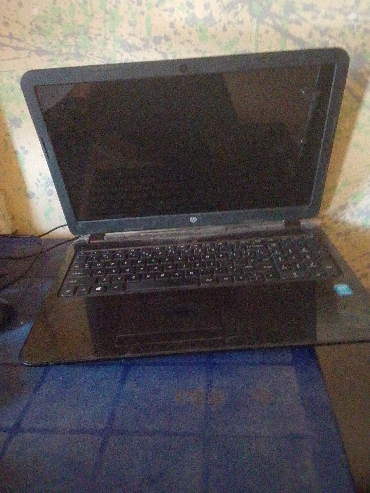 This media file shows a close-up, angled shot of an open black HP laptop. The laptop is positioned on a blue surface, possibly a table or desk, with a wall featuring a speckled, textured pattern in the background. The lighting appears to be indoors, with no indication of natural light or specific time of day. The scene is static, with no activity taking place other than the presence of the laptop.
Notable details include the HP logo clearly visible on the lid of the laptop, just above the keyboard. The keyboard itself is black with white lettering. A small, light blue sticker with a white logo, likely indicating a product feature or brand, is visible on the palm rest area of the laptop. The screen is dark, suggesting it is either turned off or displaying a dark image. There is some visible dust on the laptop, particularly on the screen bezel and around the keyboard. The overall impression is that of a functional, but perhaps older or well-used, electronic device in a domestic or office setting.
No people are visible in the frame. The context provided indicates the location is Jalingo, Nigeria, but there are no specific visual cues within the image to confirm this.