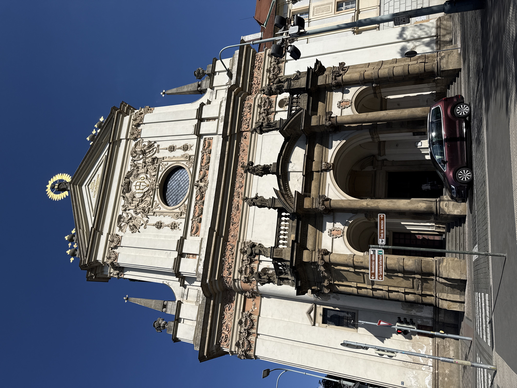 The image shows the facade of a Baroque church in Prague, Czechia, under a clear blue sky. The church is white with ornate architectural details, including columns, statues, and decorative carvings. A golden statue of a figure crowned with a halo is visible at the apex of the building. In the foreground, a street with a crosswalk leads to the church entrance, which features large arched doorways. A dark red minivan is parked in front of the church entrance. Street signs indicating directions to "Nové Městská radnice" (New Town Hall) and "Všeobecná nemocnice" (General Hospital) are present, along with traffic lights and a street lamp. The overall scene suggests a sunny day in an urban setting, likely a tourist area given the presence of directional signs.