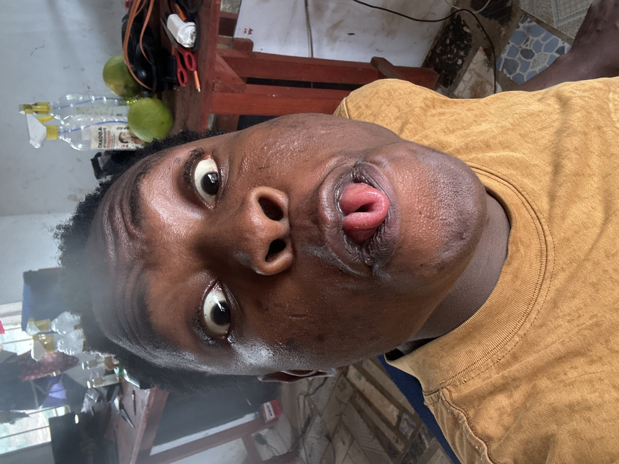 The image, oriented on its side, features a person with dark skin in a close-up, head-and-shoulders shot. The individual is making a distinctive, contorted facial expression, with their right eye wide open and staring directly at the viewer. Their left eye is also open but partially obscured by the angle. The mouth is central to the expression; the upper lip is pulled significantly upwards, revealing the upper gums, and the tongue is curled and protrudes out from between the lips in an unusual, folded manner. The person wears a mustard yellow crew-neck t-shirt.
The background to the upper left of the person's head (if the image were rotated upright) contains various household items. These include two clear plastic spray bottles, one of which has a visible label with the text "CHAOBA" and a small illustration of a face. Also present are two green, round fruits, possibly limes or small oranges, alongside red-handled scissors and tangled dark wires. This area is set against a light-colored, possibly painted, wall. To the lower right, a tiled floor with a patterned design in shades of blue, grey, and brown is visible, along with a dark wooden structure, possibly a piece of furniture. The lighting is bright and appears to be indoors. The overall impression is that of a deliberate, comical, or unusual pose.