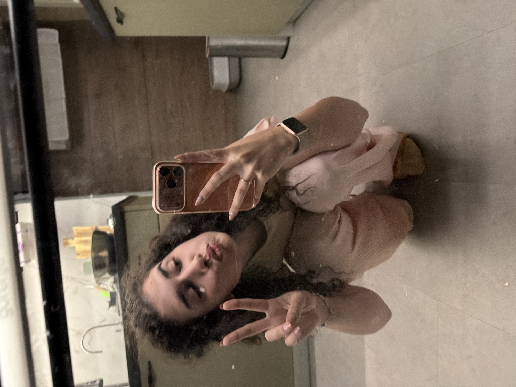In a dimly lit, domestic interior, possibly a kitchen, a young woman with curly brown hair is taking a mirror selfie. She is wearing a light pink loungewear set and is in a crouching position. Her left hand is held up, forming a peace sign, while her right hand holds a rose-gold smartphone. Her lips are pursed as if blowing a kiss. The background reveals kitchen cabinetry, a sink, and wooden cutting boards, suggesting a lived-in space. A silver trash can and a white pet litter box are visible in the distance. The flooring is a light-colored tile. The overall impression is one of casual intimacy within a private residence. Without further information, pinpointing the exact location within Sofia, Bulgaria, is not possible from the visual data alone.