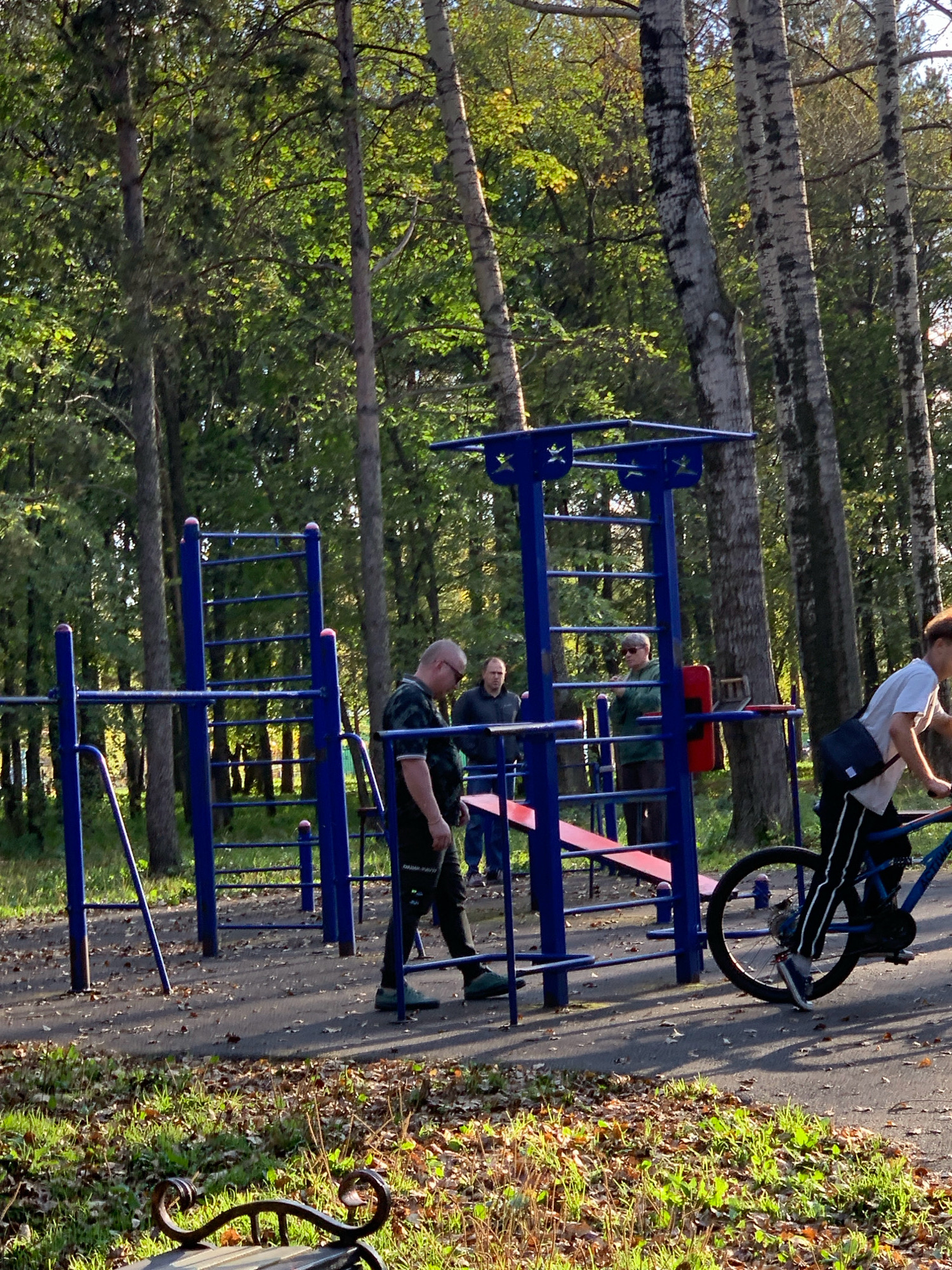 The image depicts an outdoor fitness area in Prokopyevsk, Russia, characterized by a blue metal jungle gym with various bars and ladders. Several individuals are present. In the foreground on the right, a young person on a blue bicycle is riding on a paved path, wearing a white t-shirt and black pants with white stripes. To the left of the bicycle, a man in a black shirt and black pants is standing near the fitness equipment. Further to the left, two other men are visible; one in a dark jacket and the other in a green jacket and dark pants. The environment is a park setting with tall trees, some with visible bark patterns, and dappled sunlight filtering through the leaves. The ground is covered with a mix of asphalt and scattered fallen leaves, with patches of grass and a park bench in the lower left corner.