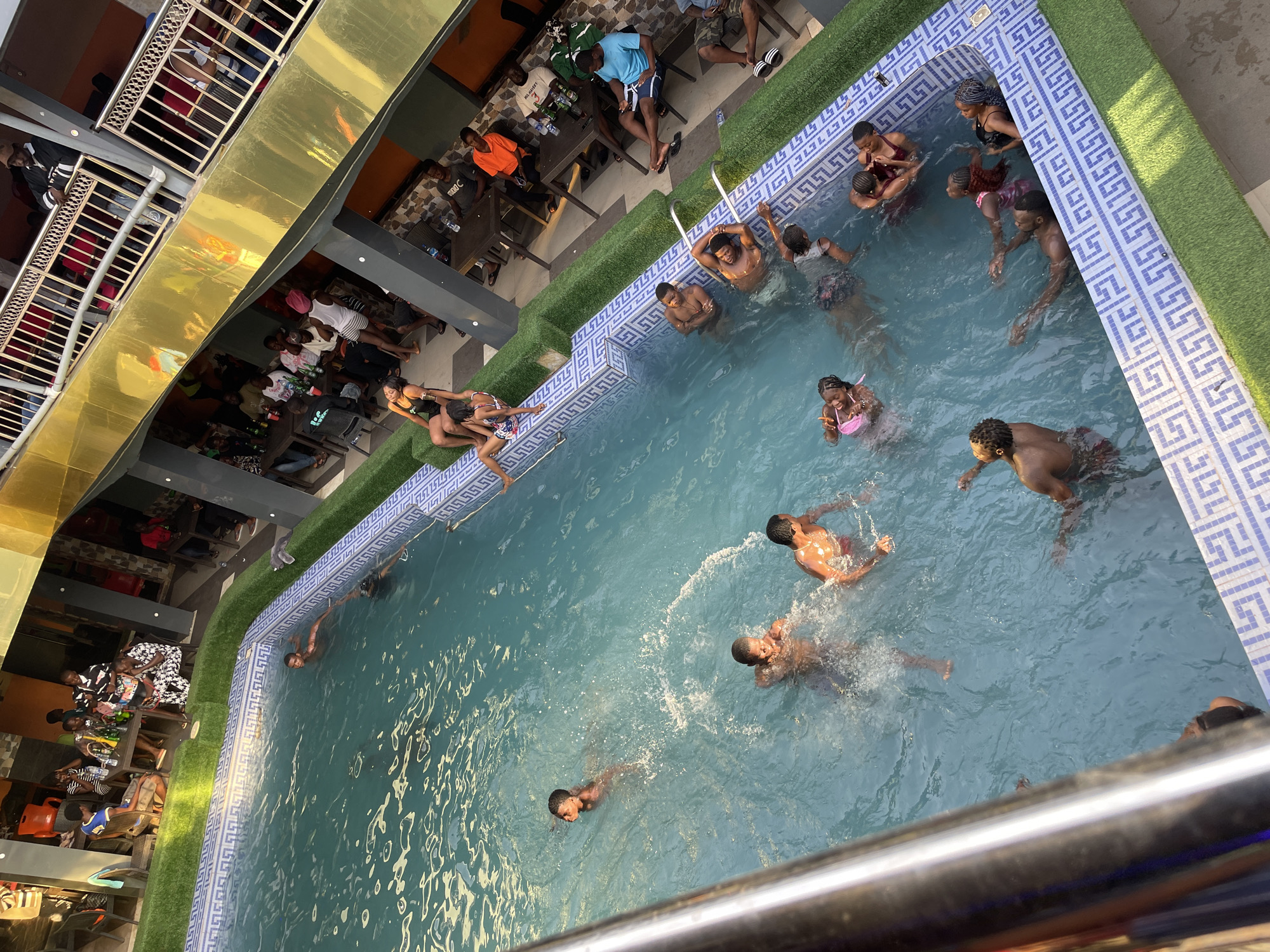 The scene depicts an elevated view of an outdoor swimming pool in Nkpor, Nigeria. Several people are swimming in the pool, while others sit at tables along the pool's edge. Some people are also on a balcony of a nearby building. The pool has a decorative blue and white tile surround, and an artificial turf border. The surrounding area appears to be an entertainment venue with multiple levels.