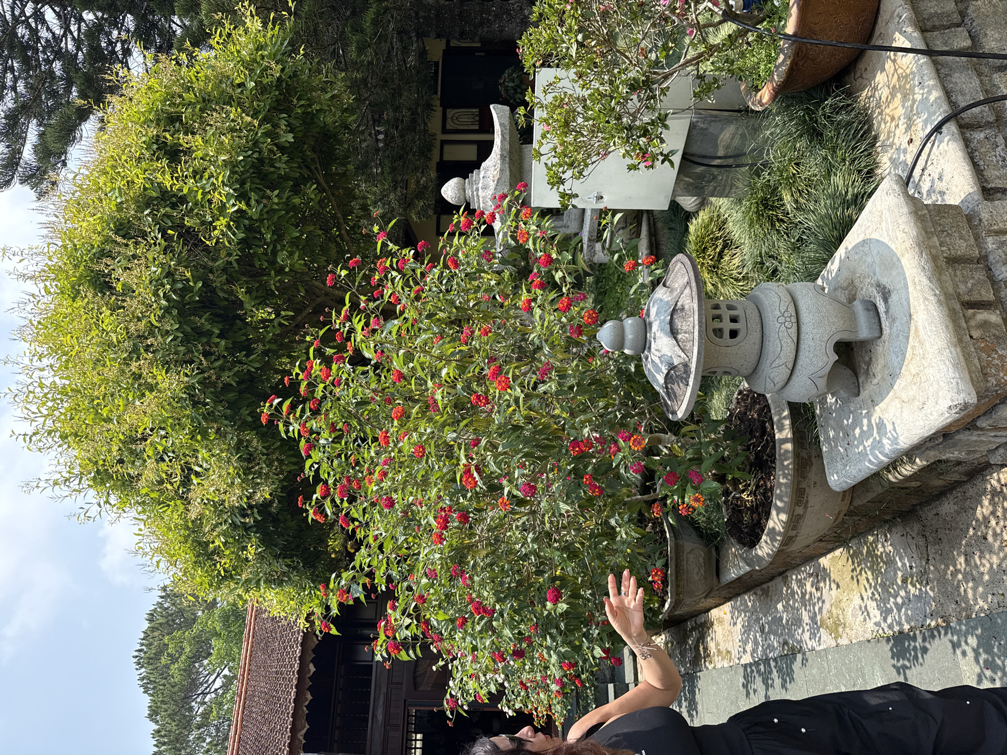 A woman with brown hair and a black top is standing outside in a garden. The garden contains trees and flowers in a stone pot. Behind the flower pot is a stone lantern. There is a second stone lantern in the background near another tree. The building is traditional architecture. The location is Da Lat, Vietnam.
