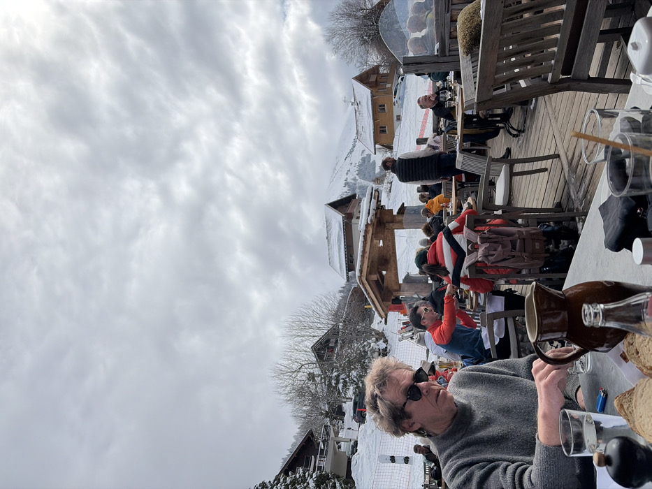 The image captures an outdoor dining terrace in La Clusaz, France, on an overcast day with a uniform layer of grey and white clouds. In the foreground, a Caucasian male with medium-length curly brown hair, wearing dark sunglasses and a grey knitted sweater, is seated at a light-colored table. His right hand is resting on the table. On the table are a clear glass of water, a blue disposable lighter, a dark shaker, slices of bread, a brown ceramic pitcher, and a clear glass bottle.
In the midground, numerous individuals are seated at wooden tables on a timber deck terrace. Patrons are dressed in various winter garments, including a person in a red and white striped jacket with a brown backpack, and another in a red top interacting with someone across a table. A male in a dark vest stands facing away from the camera, positioned among the tables. Ski equipment is visible leaned against a barrier on the left side of the terrace. A red safety net or barrier is present on the snowy slope beyond the terrace.
The background reveals a snow-covered landscape with several chalet-style buildings featuring dark roofs and light-colored walls, nestled among snow-dusted bare trees and evergreens on ascending slopes. A dark SUV is parked on a snowy path behind a chalet on the left. Distant snow-covered mountains are visible under the cloudy sky.