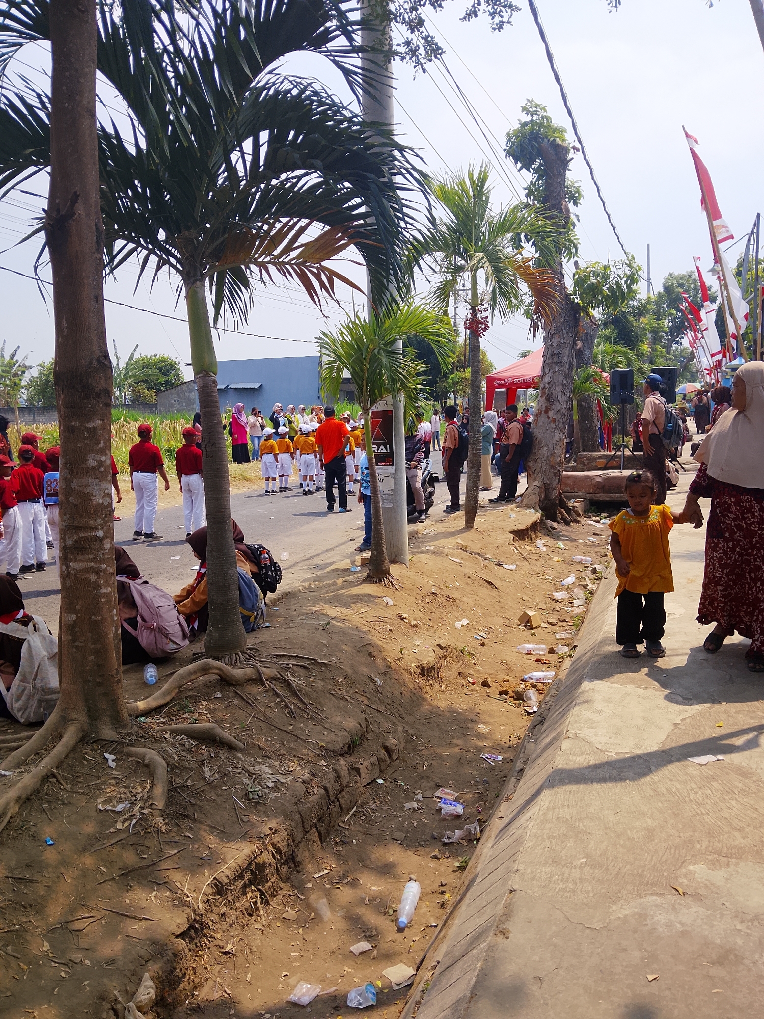 The image depicts an outdoor scene in Bangsri, Indonesia, likely on a street during a daytime event. A group of children, some wearing red and white uniforms with red caps, stand on the asphalt road. Other individuals, including adults, are also present, some watching the children. Palm trees and other vegetation frame the scene, with a blue building visible in the background. Red and white Indonesian flags are displayed along the street, suggesting a celebratory or patriotic occasion. The ground beside the road is dirt with visible roots and scattered litter, including plastic bottles and wrappers. A woman in a patterned dress and headscarf is walking on the concrete sidewalk, holding the hand of a young child wearing a yellow shirt and black pants.