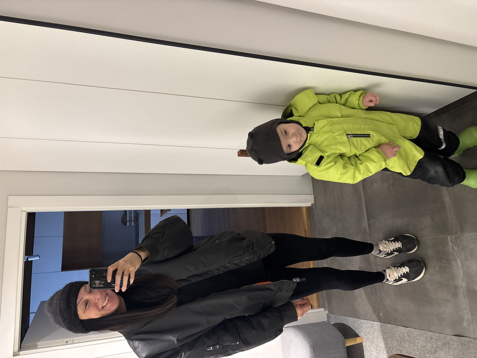 Location within Chaikovsky, Russia, is not discernible from the image.
The scene depicts an interior setting, likely an entryway, captured as a mirror reflection. An adult female, positioned on the left side, with long dark hair and wearing a dark knit beanie, a black bomber jacket with a partially visible orange lining, black leggings, grey socks, and black and white athletic shoes, smiles directly into a black smartphone held in her right hand, taking a selfie. Her left hand is by her side. To her right, a young child stands, dressed in a dark grey knit hat with two ear-like protrusions, a bright lime green and black winter jumpsuit with reflective strips, and bright lime green boots. The child's gaze is directed downwards and to the left, and they are resting against a white wall composed of vertical panels. The floor is dark grey tile. In the lower-left quadrant, a partially visible grey upholstered stool with light wooden legs is present on a grey carpeted area. Behind the adult, an open doorway reveals a room with light-colored walls, wooden flooring, and what appears to be kitchen cabinetry, illuminated by a cool, blue-tinted light source.