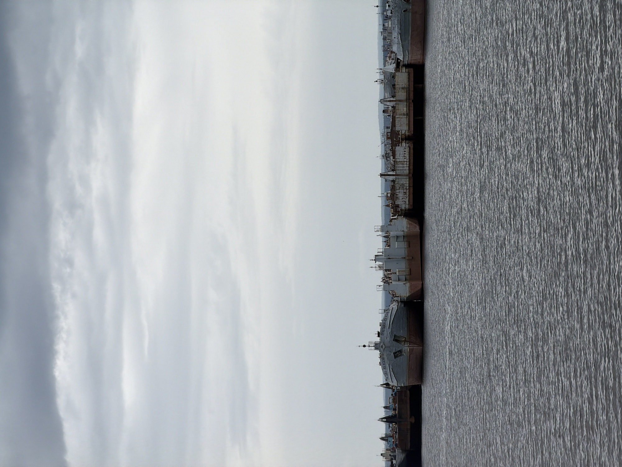 The image captures a serene, somewhat somber scene featuring numerous old ships or barges docked along a body of water under an overcast sky. The photograph is taken from the water, looking towards the shore where the vessels are moored in a long line. The water surface shows subtle ripples, indicating calm conditions.
The main subjects are the vessels themselves, which appear to be utilitarian river barges or cargo ships, many showing signs of age, rust, and disuse. Their structures are generally flat-decked with small superstructures, masts, and various equipment, suggesting they might be industrial or transport vessels. No people are visible, emphasizing the quiet, perhaps abandoned, nature of the scene.
The sky occupies the upper portion of the image, displaying a mostly uniform, soft gray light characteristic of an overcast day. There are some lighter and darker patches of clouds, but no direct sunlight is evident. The overall color palette is muted, dominated by grays and desaturated browns from the ships' hulls.
Notable details include Cyrillic text visible on the bow of one of the ships. It reads "ПРИСТАНОВКА ЗДЕСЬ ЗАПРЕЩЕНА", which translates to "MOORING IS FORBIDDEN HERE". Below this, another line of text is partially visible, appearing to say "ПРИСТАНОВКА ЗДЕСЬ" again, followed by "ЗАПРЕЩЕНА". Further down on the hull, partially obscured, is "ЧАЙКОВСКИЙ", which corresponds to the provided location, Chaikovsky, Russia, a town on the Kama River, implying these are river vessels. The presence of this text on the ships suggests the location is indeed Russia, and specifically Chaikovsky, making the vessels likely moored in a restricted area or a lay-up facility.