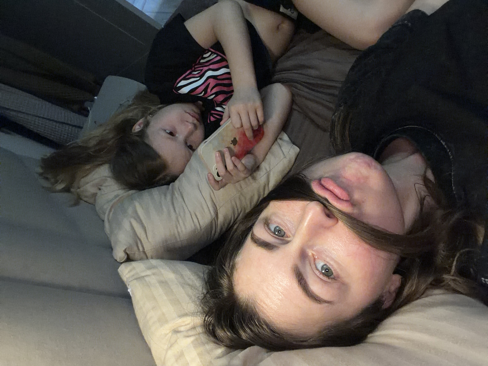 Two individuals are lying on a bed. The individual closer to the camera is an adult woman with fair skin, light eyes, and dark hair. She is lying on a light-colored pillow. A younger individual, presumably a child, is partially visible behind her, also lying on a pillow, and is holding a cell phone. The child is wearing a black shirt with a pink and white design. The bed has a brown-colored comforter. There is no evidence to suggest a location in Dubovaya, Russia. The image is a candid photograph.