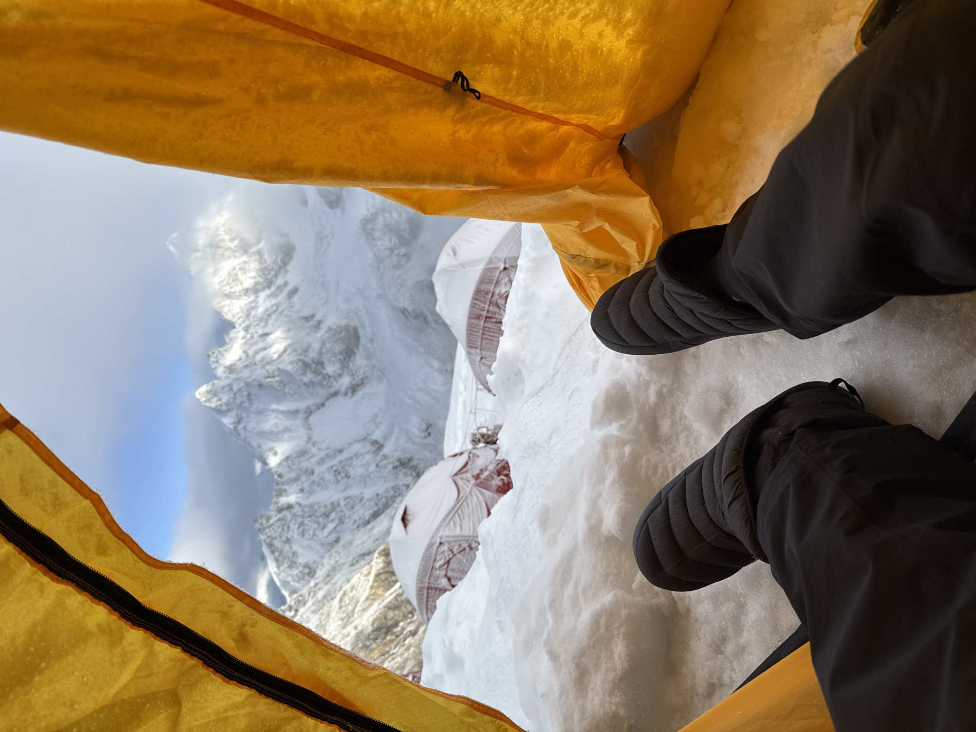 The image is taken from inside a yellow tent, looking out at a snowy mountain landscape. In the foreground, a pair of black quilted slippers and black pants are visible, suggesting a person is inside the tent. Outside the tent, several snow-covered red and white tents are partially buried in the snow. Beyond the tents, a majestic, snow-capped mountain range dominates the background, with some peaks shrouded in clouds. The sky is blue with scattered clouds. The location is identified as Samagaun, Nepal, a village situated in the Gorkha District of Nepal, near the Tibetan border and at the foot of Mount Manaslu, the world's eighth highest mountain.