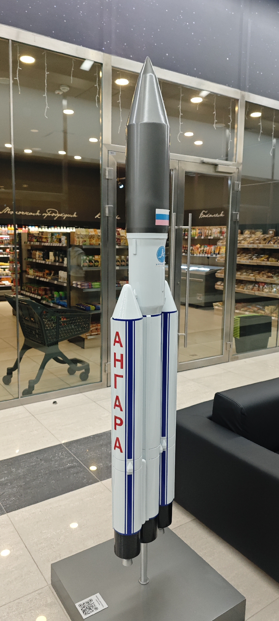 Here is a description of the image:
A model of the Angara rocket is situated inside a retail establishment. The model is predominantly white with blue stripes and red Cyrillic lettering spelling "АНГАРА." A small Russian flag is affixed near the top of the rocket body, beneath a grey section. The rocket stands on a grey base which also has a QR code sticker on it.
In the background, aisles of goods are visible behind glass doors and walls. A shopping cart can be seen to the left of the rocket, and a black couch is situated on the right. Overhead lighting is reflected in the glass, and the image is taken indoors. The location is the city of Estosadok, Russia.