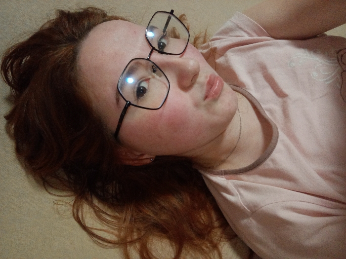 A close-up photograph depicts a young individual with reddish-brown hair, lying on a light, neutral-colored surface, positioned diagonally within the frame. The subject wears a light pink t-shirt with a darker pink collar trim and black, hexagonal-framed eyeglasses. A thin silver-colored chain is visible around their neck. The subject's lips are slightly parted and pursed in a pouting expression, with a glossy appearance. Both eyes are visible through the eyeglasses, gazing towards the camera, with light reflections present on the lenses. The cheeks show a flushed complexion. The surrounding environment is an indistinct, uniform light-beige texture. This scene is located in Старый Шагирт, Russia.