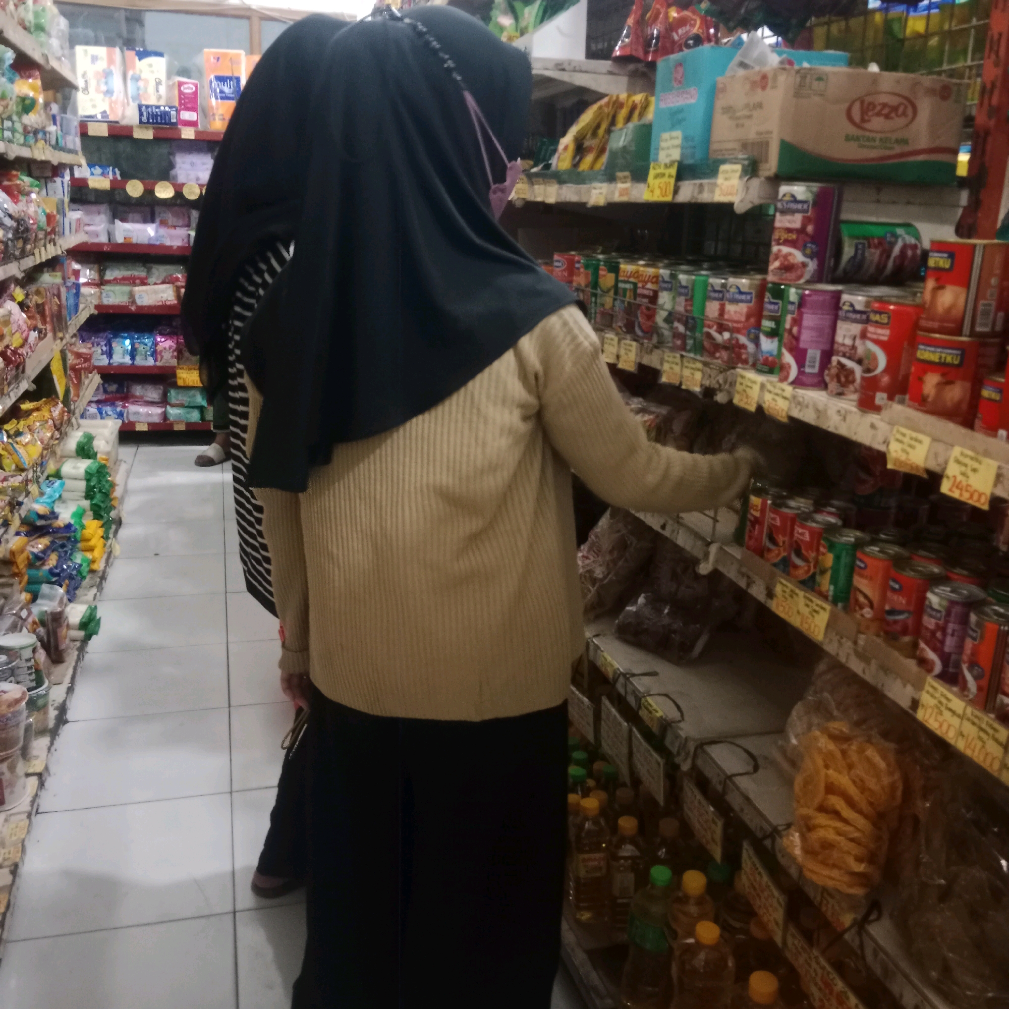 The image depicts an indoor scene, likely a grocery store or convenience store in Tegowanu Wetan, Indonesia. Two individuals are visible, both wearing hijabs. One person, in the foreground, is wearing a tan sweater and black pants, and appears to be reaching for items on a shelf. The other person, partially obscured by the first, is wearing a black hijab and a striped garment. The shelves are stocked with various consumer goods, including canned foods, packaged snacks, and bottled liquids, some with visible price tags. The flooring is tiled and appears to be light-colored. The overall lighting suggests an indoor environment, possibly with artificial lighting.