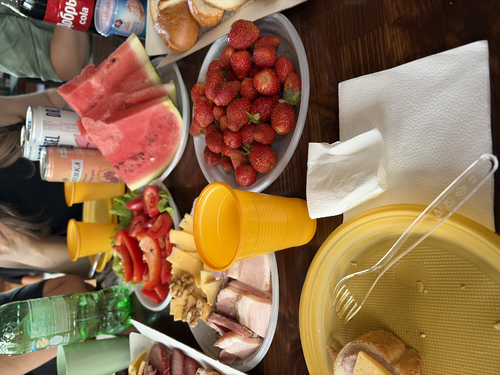 A close-up, overhead shot captures a wooden table laden with picnic food and drinks, likely in Chaikovsky, Russia. In the foreground, a yellow plastic plate holds a half-eaten sandwich and a clear plastic fork. To its right, a white paper napkin is folded. A plate of fresh strawberries sits to the right of a white plastic plate containing sliced bacon and cheese with walnuts. Behind that, a slice of watermelon is on a white plate. Further back, there are several canned drinks, including a Coca-Cola bottle with a Cyrillic label. Two yellow plastic cups are also visible, along with a green plastic cup and a green plastic bottle. In the background, blurry figures of people can be seen. The overall scene suggests a casual outdoor gathering.