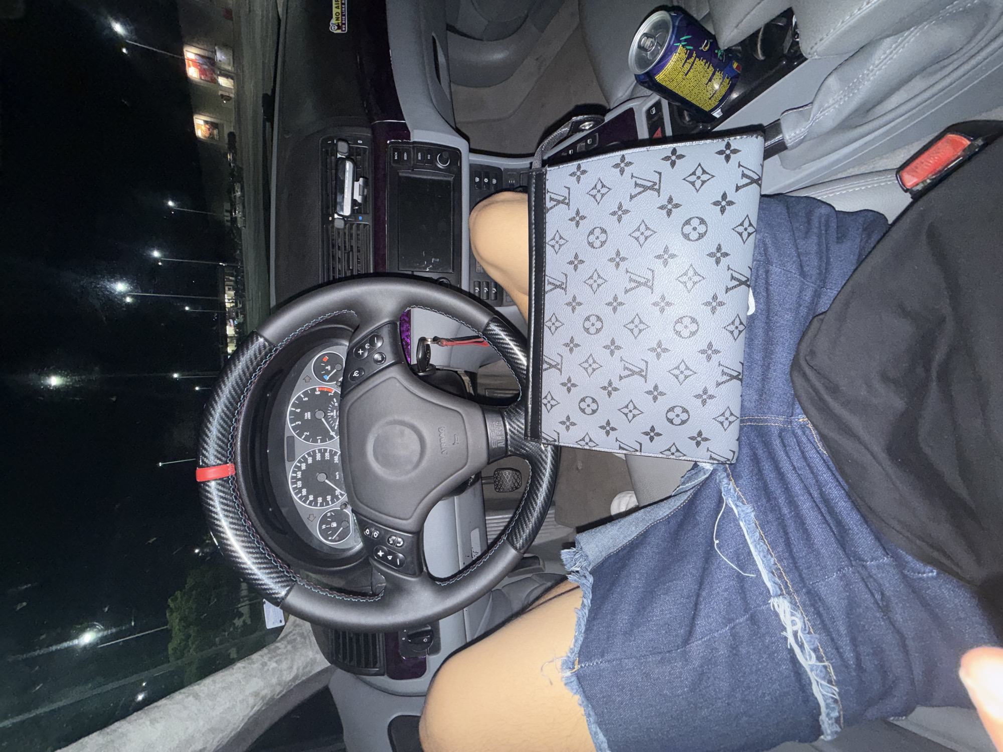 The image shows an interior view of a car at night. A person is visible, wearing denim shorts and a black top, sitting in the driver's seat. A Louis Vuitton bag rests on their lap, partially obscuring their legs and shorts. The car's steering wheel and dashboard are prominently visible, along with a beverage can in the center console area. The background appears to be a nighttime street scene. There is no information available to establish if this photo was taken in Burgas, Bulgaria.