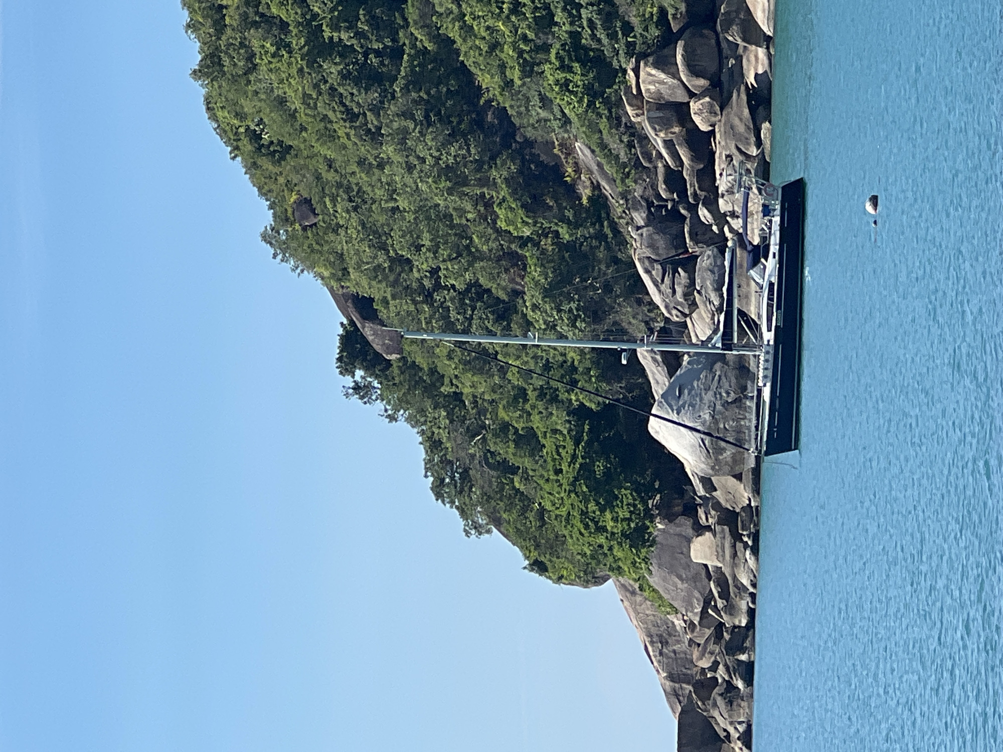 A dark blue sailboat with a white stripe and a white cabin is anchored in turquoise water near a rocky, tree-covered island. The island features large boulders and lush green vegetation under a clear blue sky. The boat has a tall mast with rigging and appears to have a few people on deck, though they are not clearly visible. A small white buoy is floating in the water to the right of the boat. The scene is located in Baan Chaweng Noi, Thailand.