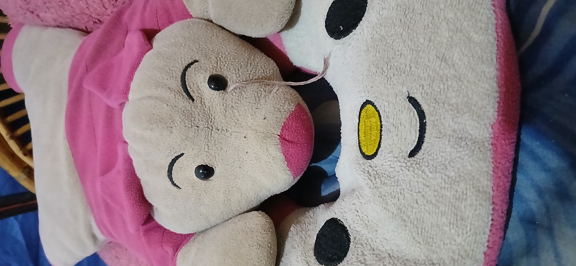 The image shows a close-up of a stuffed animal, likely a pillow or a toy, with a pink and cream color scheme. The stuffed animal has a face with two black eyes, arched black eyebrows, and a pink snout. One of the eyes has a loose pink thread hanging from it, resembling a tear. Below the face, there is a white section with embroidered features of a face, including two black oval shapes representing eyes, a yellow oval shape with embroidered texture, and a curved black line for a mouth. The stuffed animal is resting on a blue and striped surface, possibly a bed or sofa, with some wooden elements visible in the background. The lighting suggests an indoor setting. There is no discernible information within the image to indicate its location in Samarinda, Indonesia.