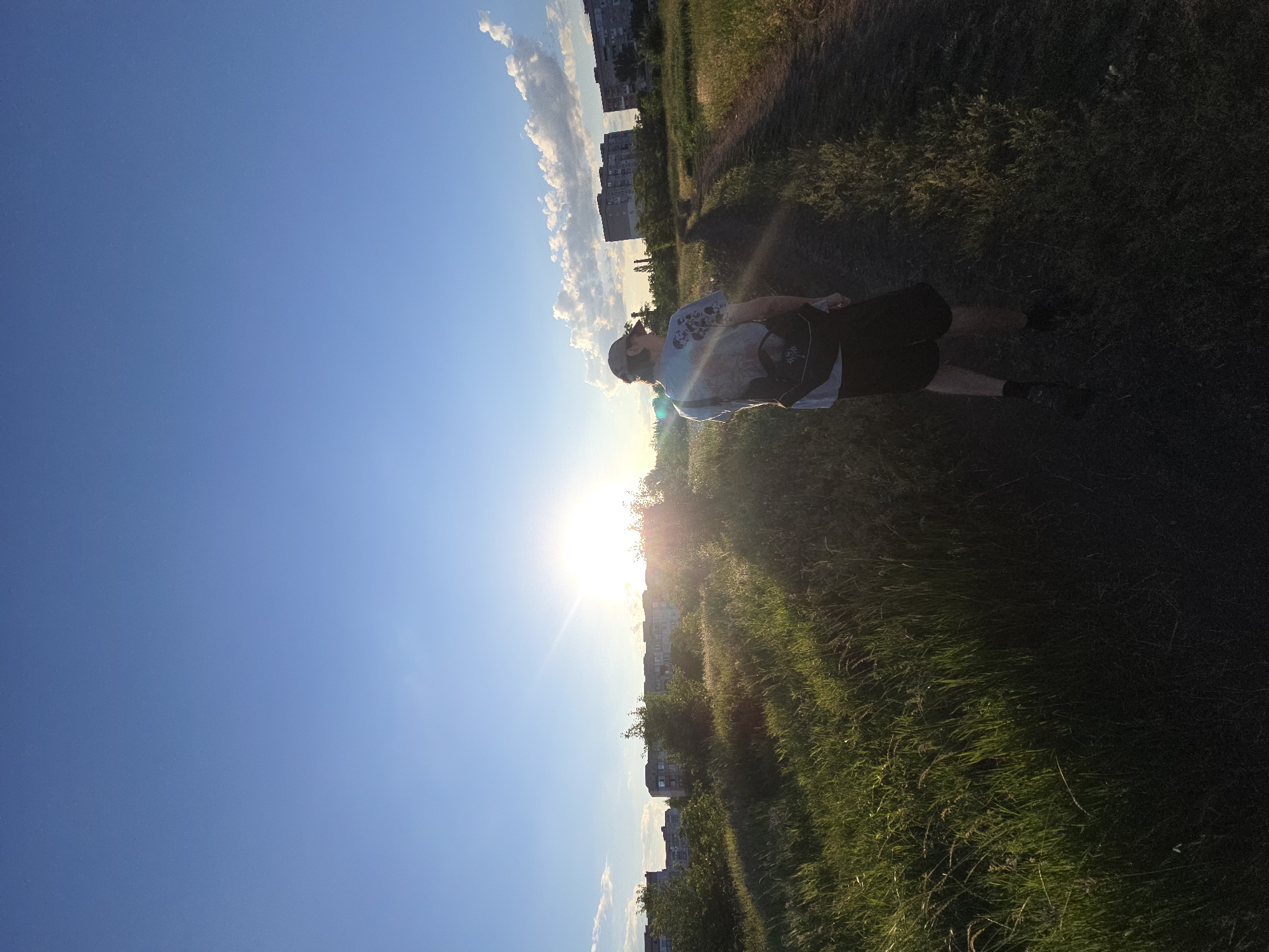 A lone figure is walking away from the camera down a dirt path, with tall green grass on their left and a line of apartment buildings in the distance on the right. The sun is low on the horizon, casting a bright glare and long shadows. The sky is clear and blue with a few wispy clouds. The person is wearing a baseball cap, a graphic t-shirt, and shorts, with a backpack slung over one shoulder. The overall scene depicts a warm, late afternoon or early evening in an urban fringe environment.