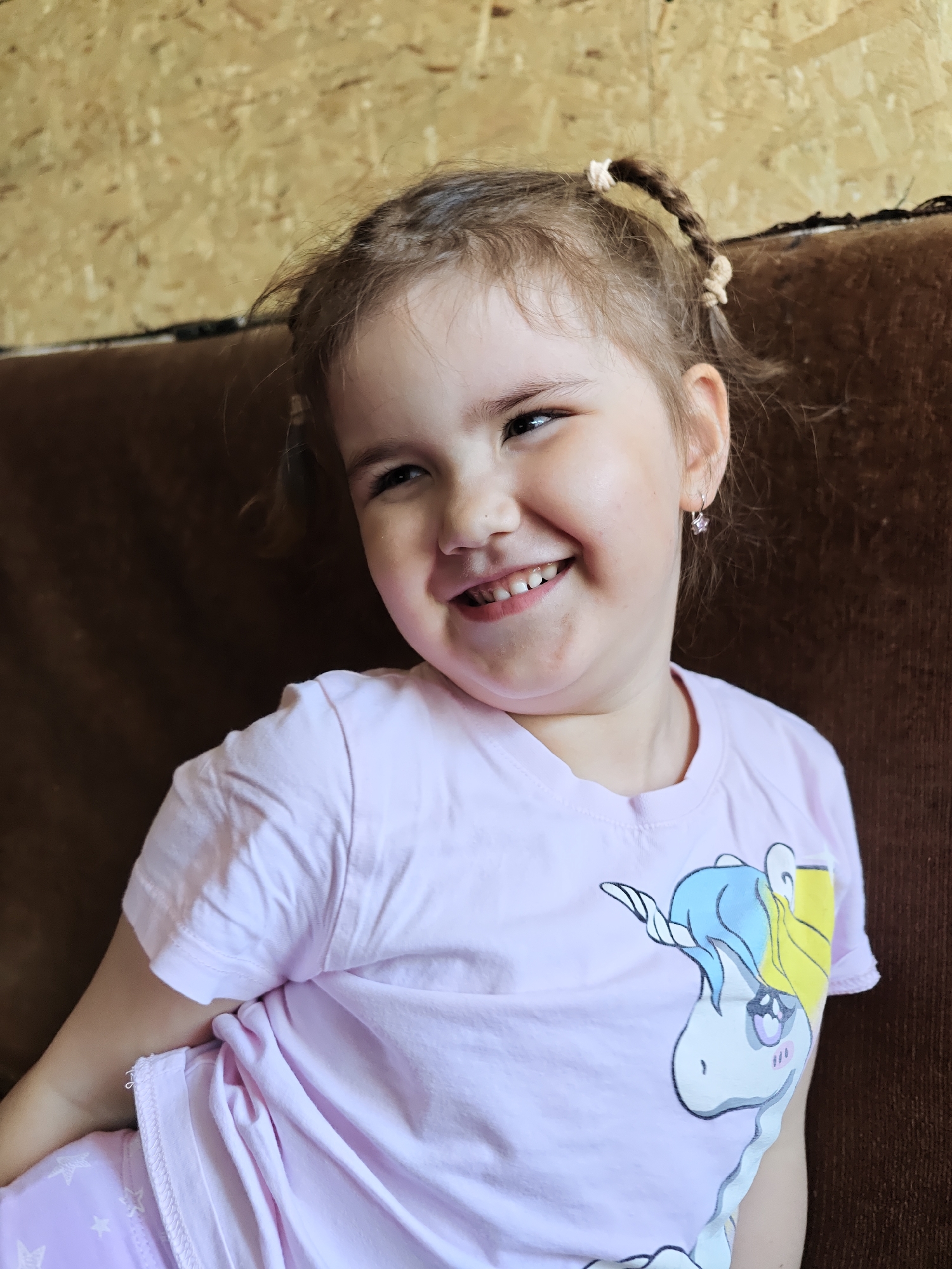A young girl is seated indoors, likely on a brown sofa. She is wearing a pink T-shirt with a unicorn graphic and matching pink pajamas. Her light brown hair is styled in two small pigtails secured with light-colored elastics. She is smiling, her face turned slightly to the side. She is wearing small earrings. The background features a light brown wall. The location is Malyi Bukor, Russia.