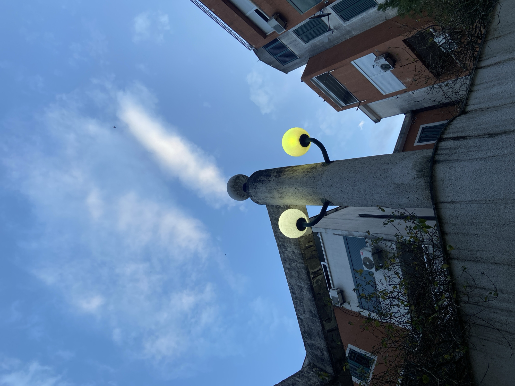 The image shows a low-angle view of a street scene. In the foreground is a weathered, concrete structure with two globe-shaped streetlights attached. The upper streetlight is illuminated with a yellow glow, while the lower one emits a cooler, white light. The structure appears to be part of a building or wall.
Beyond the structure, the sky is visible with scattered clouds. A small aircraft can be seen in the distance.
Adjacent to the right side of the concrete structure are buildings with a mix of white and brown exteriors. Some windows have dark blue shutters. Air conditioning units are visible on the walls of the buildings. Vines and foliage are growing on the structure. The location is an urban setting. The city and exact location are unknown.