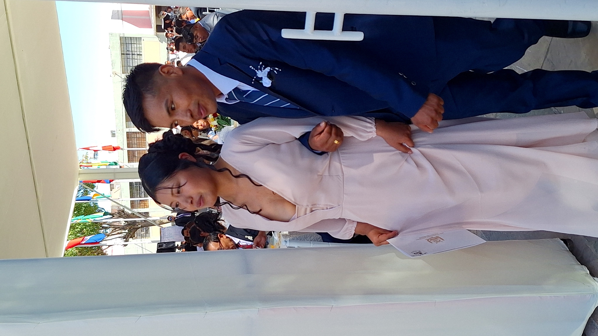 The image shows a couple at an event in Cerro Colorado, Peru. The woman is wearing a light-colored, long-sleeved dress, while the man is in a dark blue suit with a patterned tie and a white flower boutonniere. They are standing under a white tent-like structure. In the background are other people and buildings, with colorful flags visible. The woman is holding a document with a crest on it.