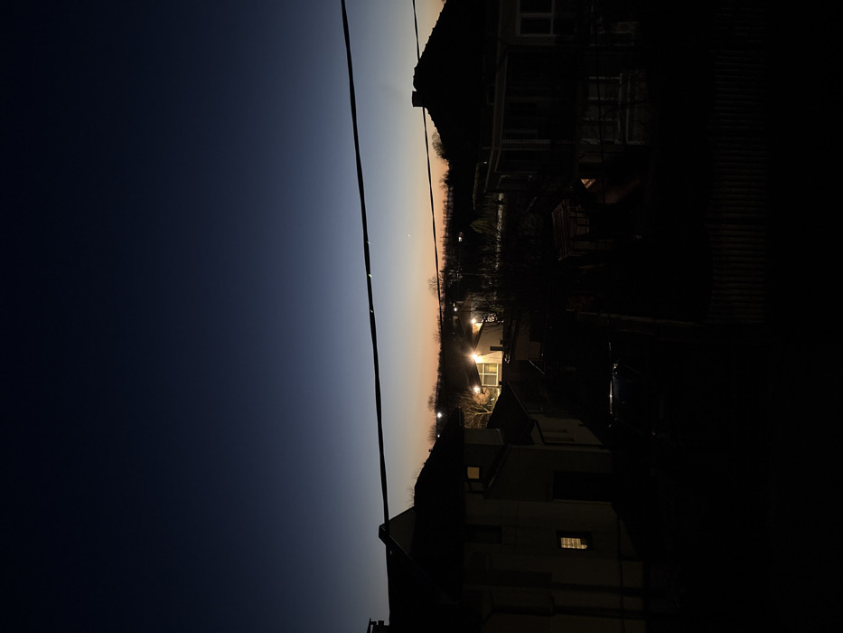 The image depicts a residential area in Stefan Karadja, Bulgaria, during twilight or early night. The sky transitions from a deep indigo at the top to a muted orange-yellow glow at the horizon, where a single, faint celestial body is discernible. A thick, dark power line spans horizontally across the upper-middle portion of the frame, exhibiting several reflective points.
In the foreground and mid-ground, multiple detached residential houses are visible, primarily as dark silhouettes against the ambient light. On the left, a light-colored multi-story house features a single illuminated window on its side, emitting a warm yellow light. On the right, a darker house with a prominent chimney and a distinctive roofline is also silhouetted. Further into the mid-ground, additional houses are present, with several artificial lights illuminating their exteriors or adjacent areas, most notably a house in the central background with bright facade lighting. Deciduous trees and shrubs, devoid of leaves, are scattered throughout the scene. A dark, low fence marks the bottom edge of the foreground. A dark vehicle is partially visible between two houses in the mid-ground. No individuals are present, and no overt actions or interactions are depicted.